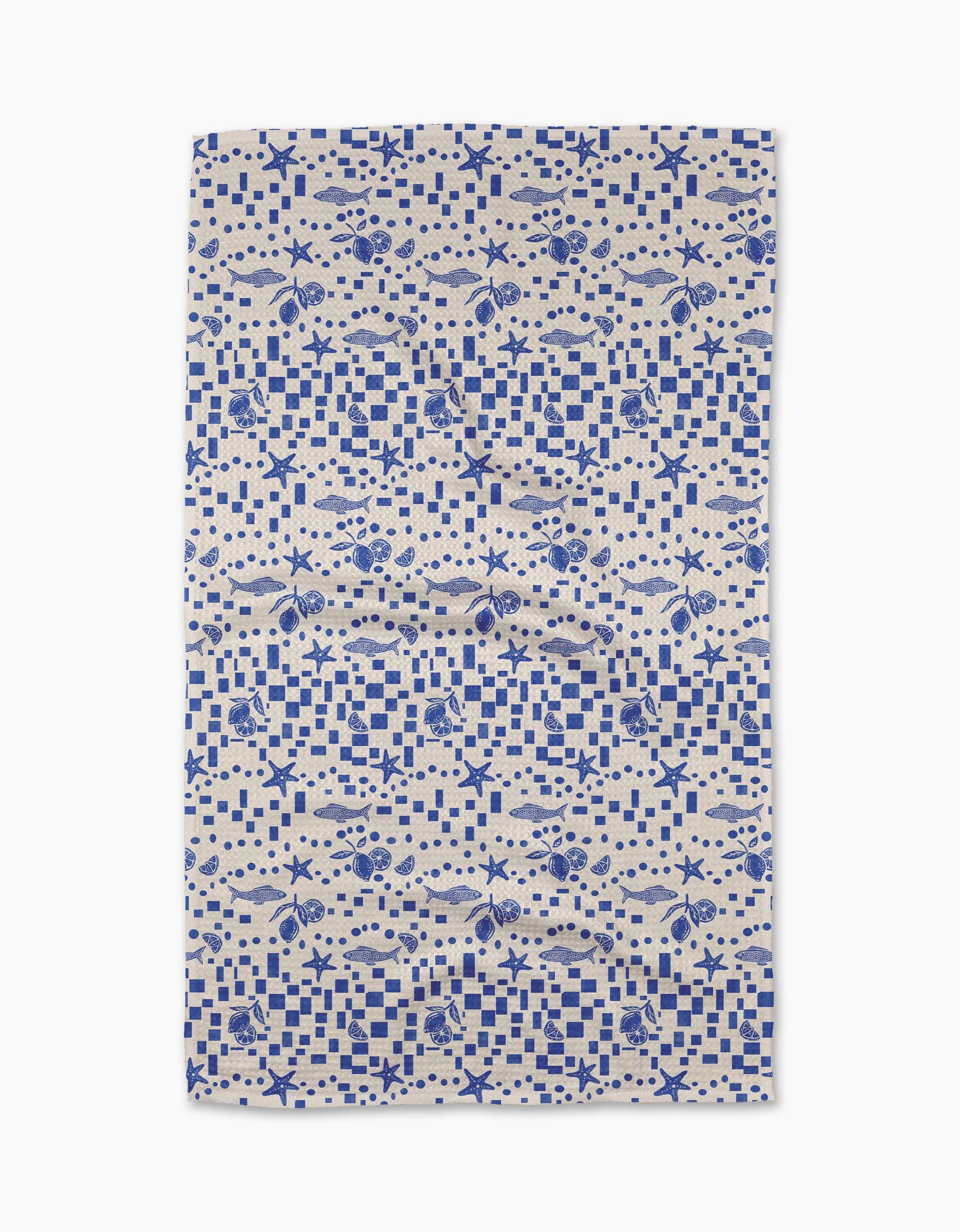 The Santorini Tile Tea Towel by Geometry is a rectangular beige towel with a blue nautical pattern of fish, seashells, starfish, and checkerboard designs. Made from recycled materials, it’s shown flat on a white background.