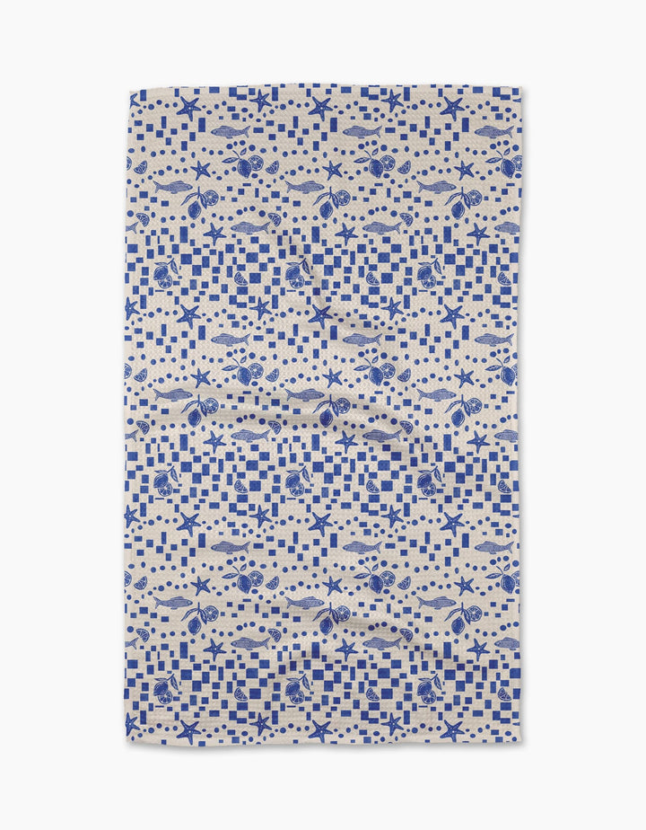The Santorini Tile Tea Towel by Geometry is a rectangular beige towel with a blue nautical pattern of fish, seashells, starfish, and checkerboard designs. Made from recycled materials, it’s shown flat on a white background.