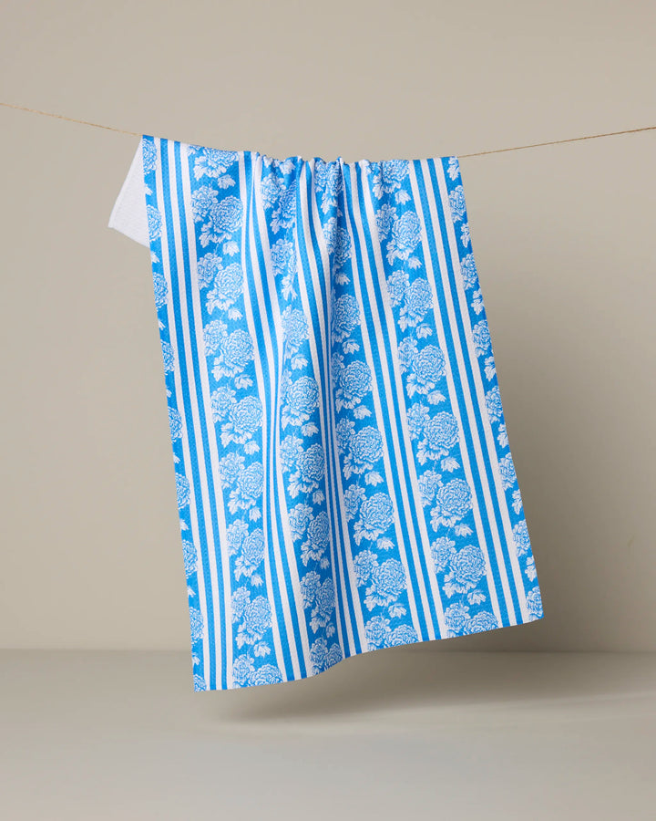 The Santorini tea towel by Geometry, featuring blue and white stripes with floral patterns, hangs on a clothesline against a neutral background.