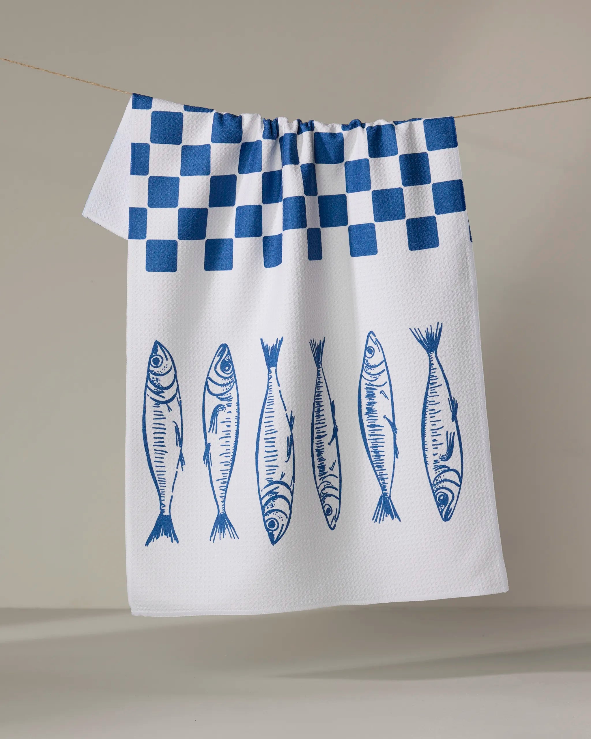 The Sardine Check Blue tea towel by Hufton Studio dries quickly and features a blue checkered top with six blue illustrated fish lined up vertically on the lower half.