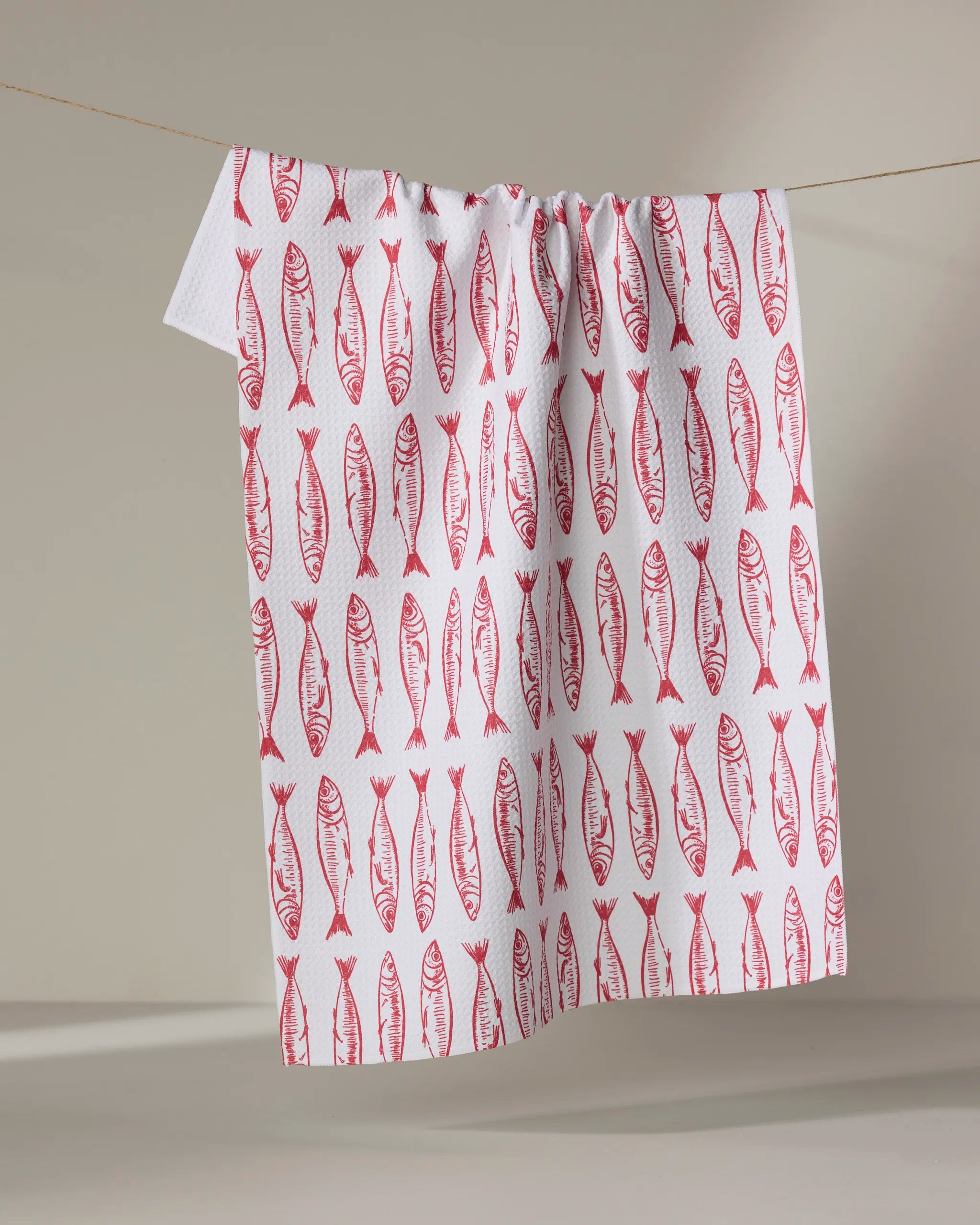 The "Sardine Stripes" tea towel by Hufton Studio features a repeating red fish pattern and is shown hanging on a clothesline against a neutral background.