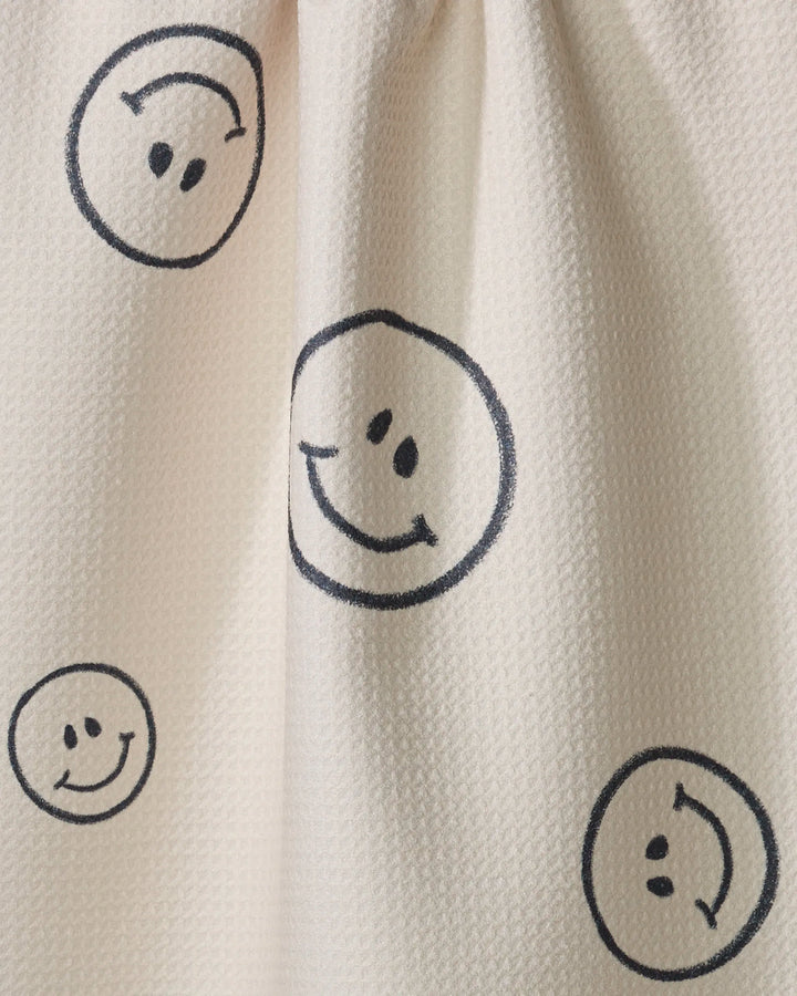 The Say Cheese tea towel by Kelsi Fullmer is cream-colored with a textured feel, decorated with scattered hand-drawn black smiley faces in various sizes, and made from recycled materials.