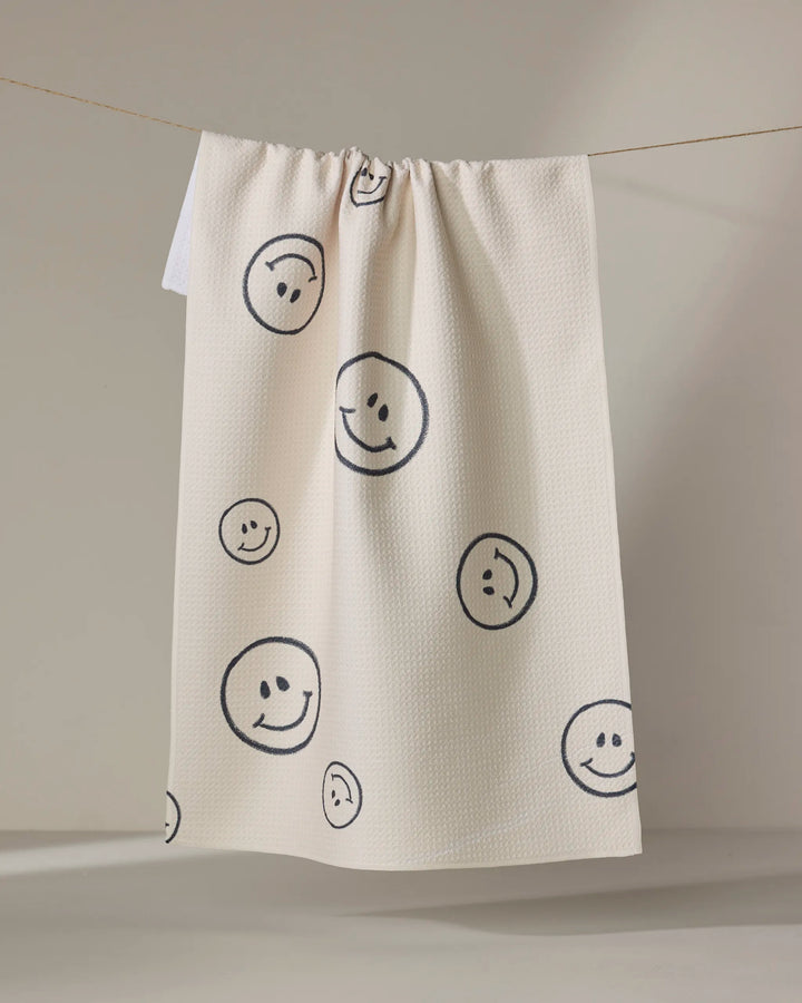 The Say Cheese tea towel by Kelsi Fullmer is cream-colored, made from recycled materials, and features black outlined smiley faces of various sizes on its textured, quick-drying surface.