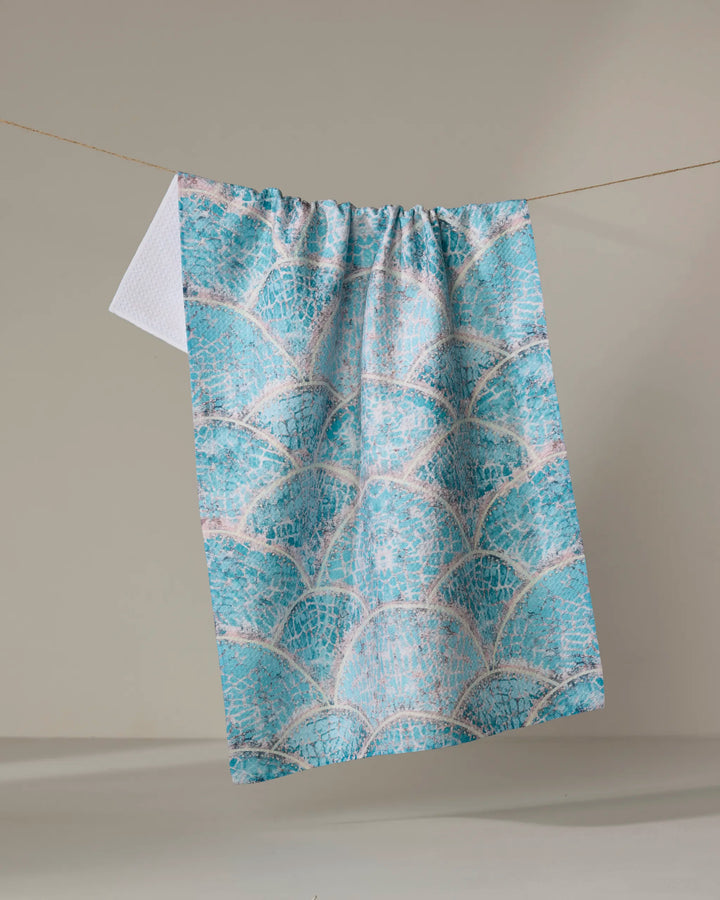 The Taryn Brennan "Scallop Hills" kitchen towel, featuring a blue and white scalloped pattern and made from recycled materials, hangs from a clothesline against a neutral background.