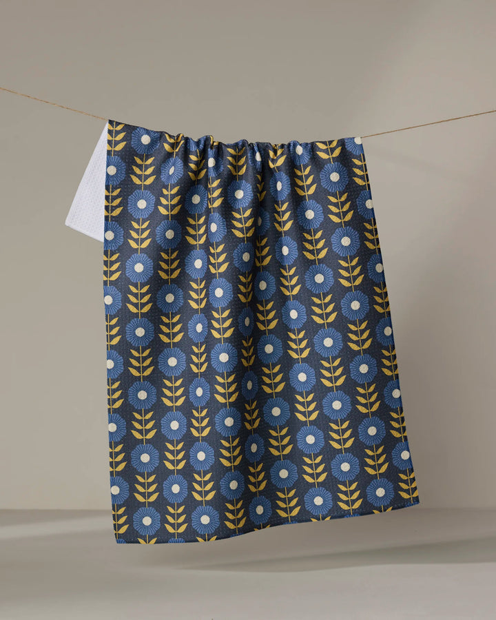 The Scandi Floral tea towel by Megan Kampa hangs on a clothesline, displaying a blue background with yellow leafy stems and white circular flowers in a repeating pattern.