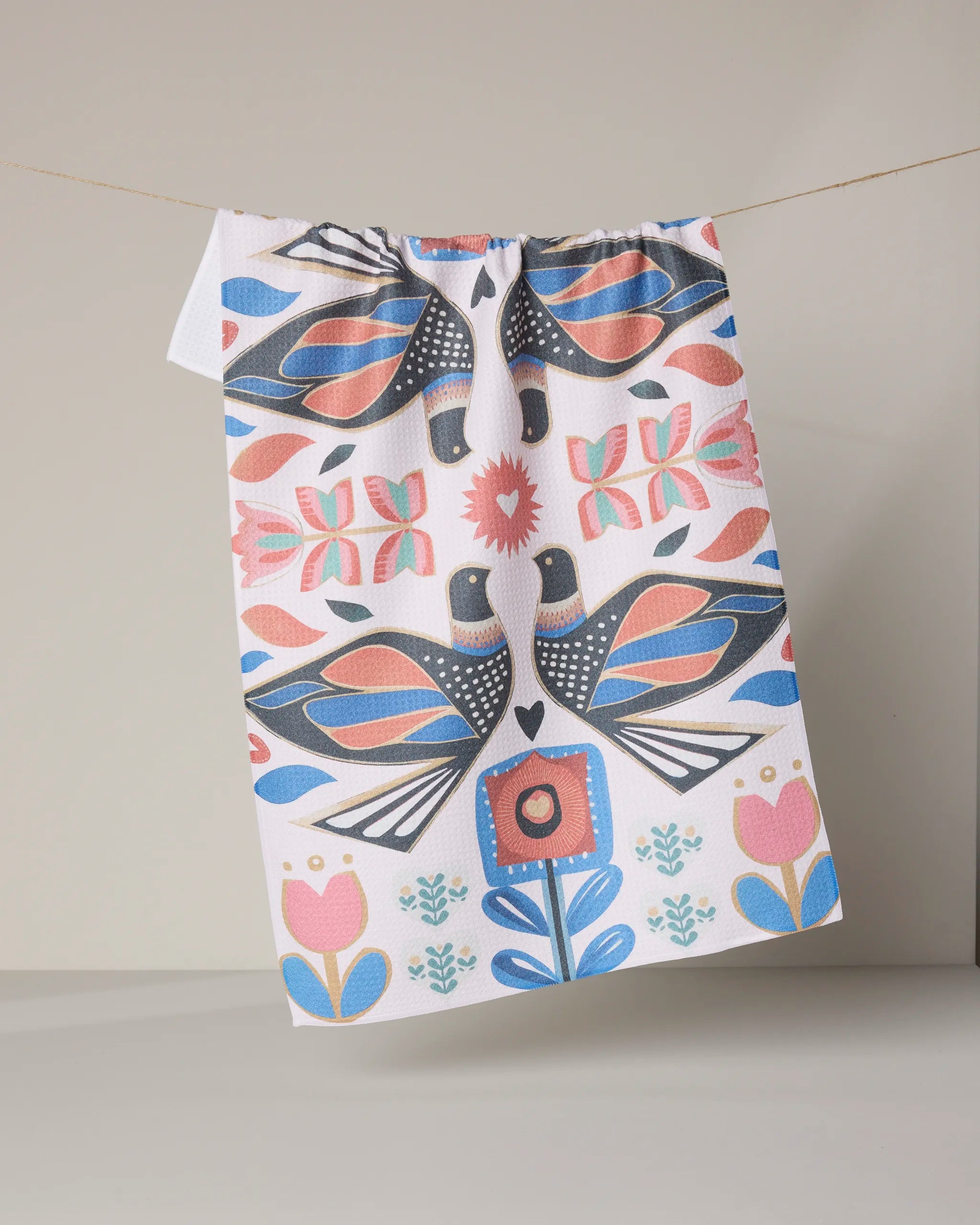 The Geometry “Scandi Lovebirds” kitchen tea towel, made from recycled materials, displays a vibrant folk art design with symmetrical birds, flowers, and hearts in blue, pink, and coral tones. It hangs on a string against a neutral background.