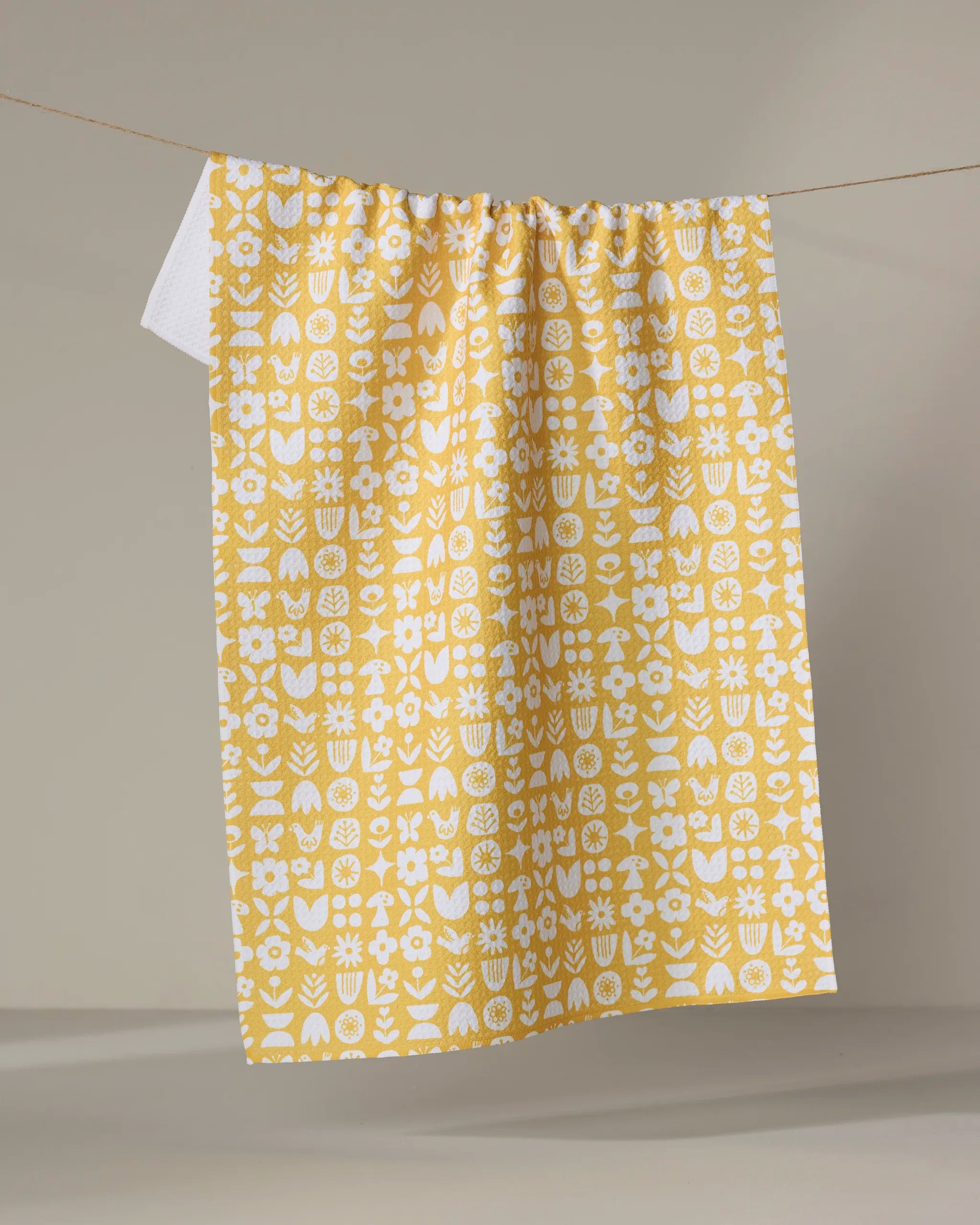 The Kayla Ann "Scandi Spring" yellow tea towel, made from recycled materials and adorned with a playful white floral pattern, hangs over a clothesline against a neutral background.