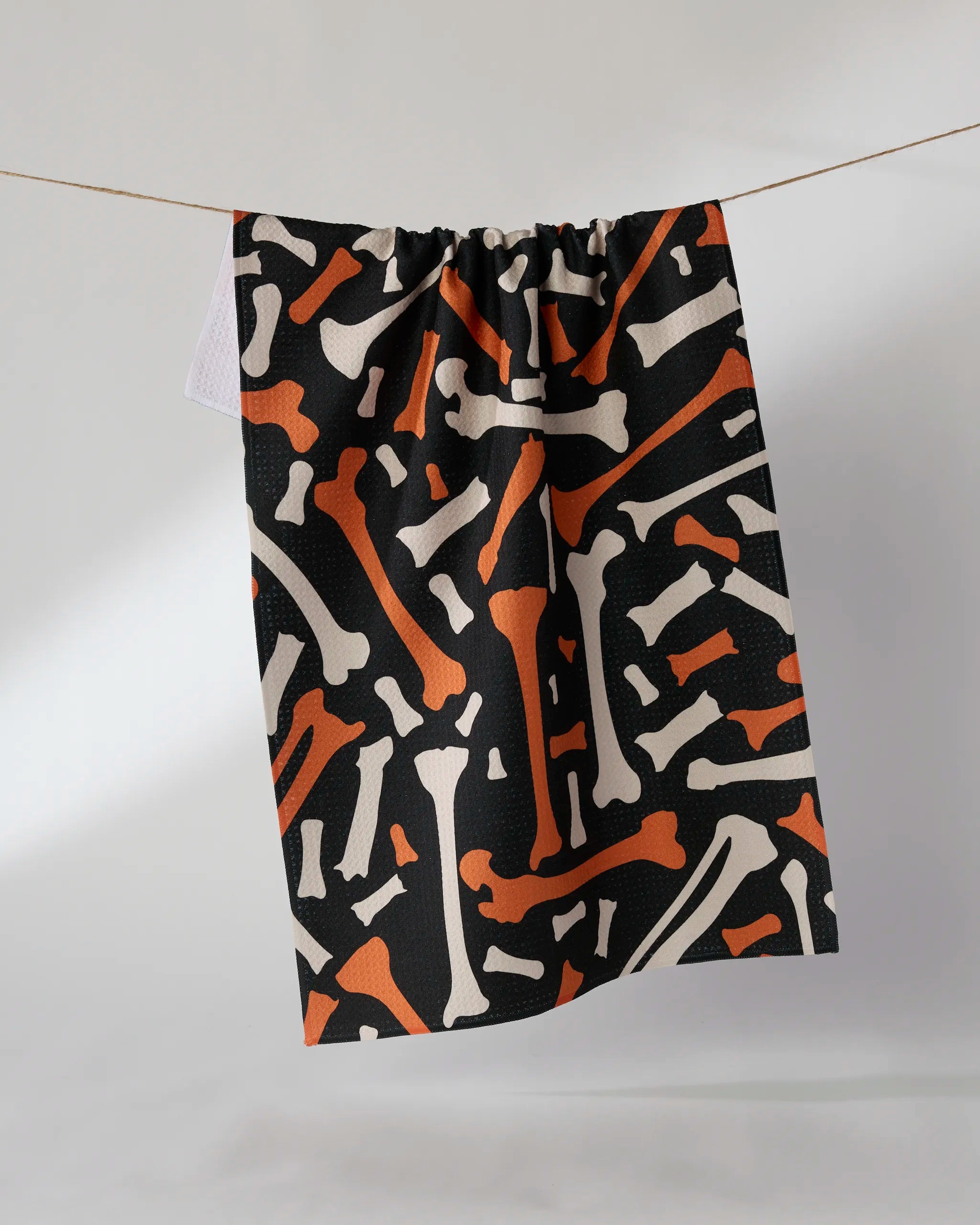 The Geometry Scatter Bones tea towel, ultra-absorbent with an orange and white bone pattern on black, hangs on a clothesline against a plain light wall.