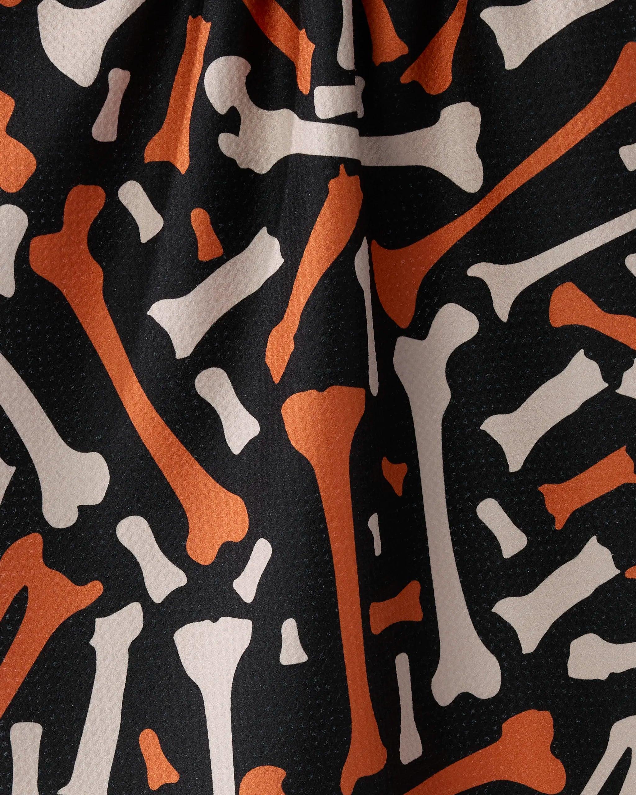 The Geometry Scatter Bones fabric features orange and white cartoon bones scattered on a black background, made from recycled materials with small textured dots and visible folds.