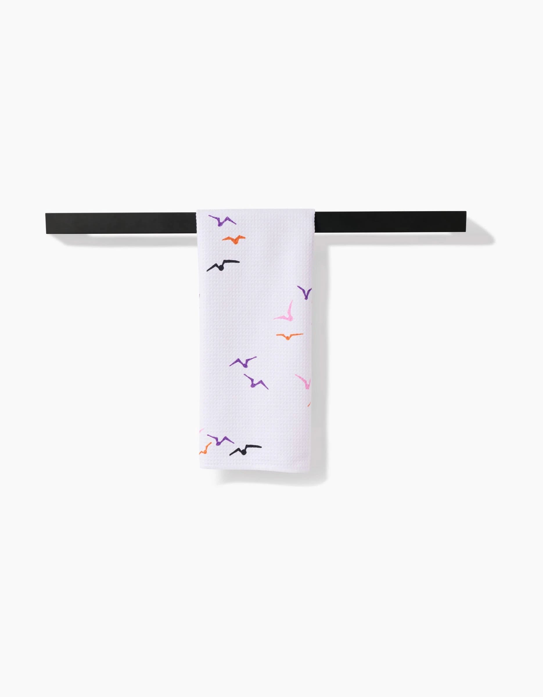 The Scattered Seagulls tea towel by Ania Zwara Art, made from recycled materials, features white textured fabric with orange, purple, and black bird silhouettes draped over a black bar on a plain white background.