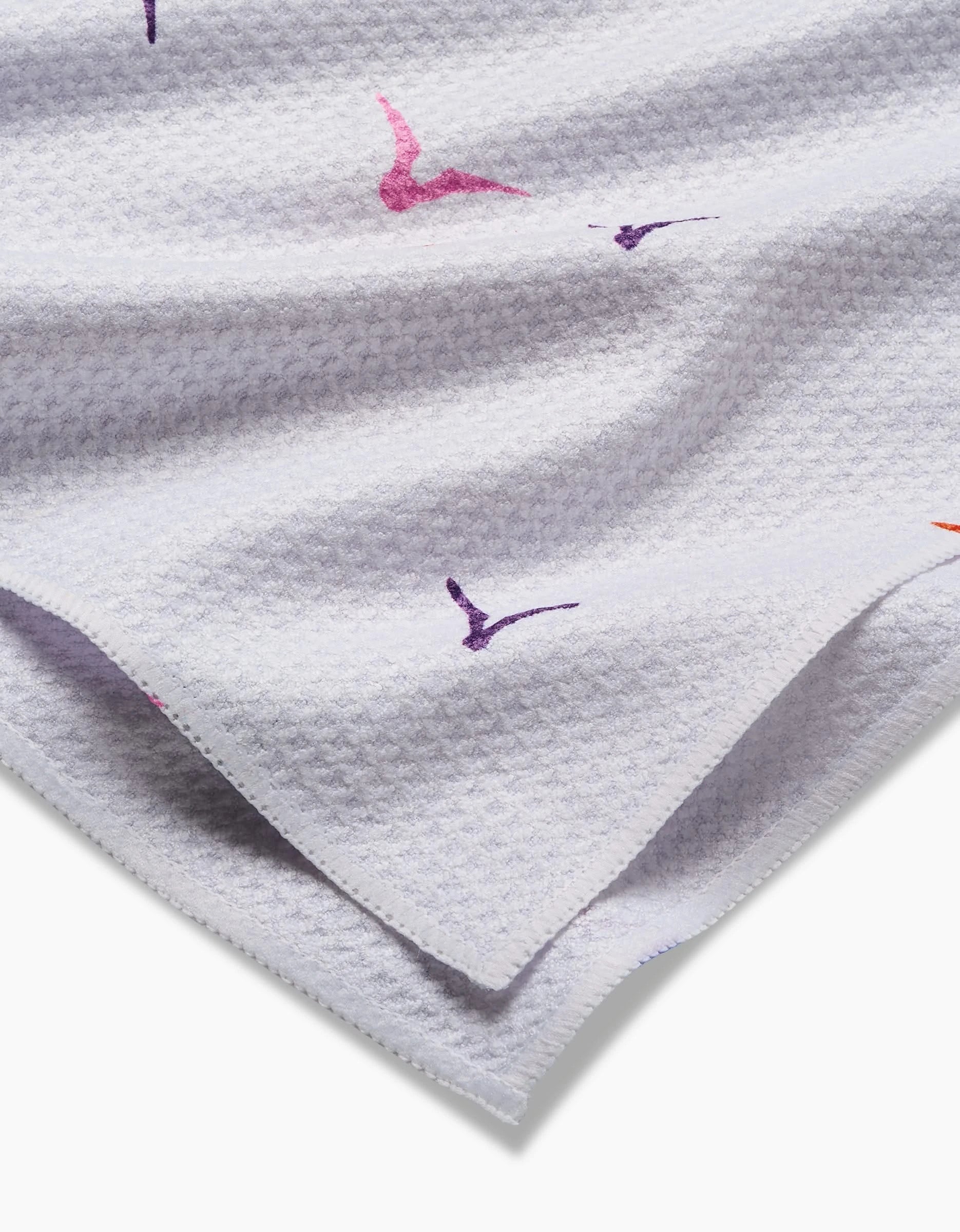 A close-up of the Scattered Seagulls tea towel by Ania Zwara Art shows a white waffle weave texture adorned with pink, purple, and orange bird silhouettes scattered across its quick-drying surface.