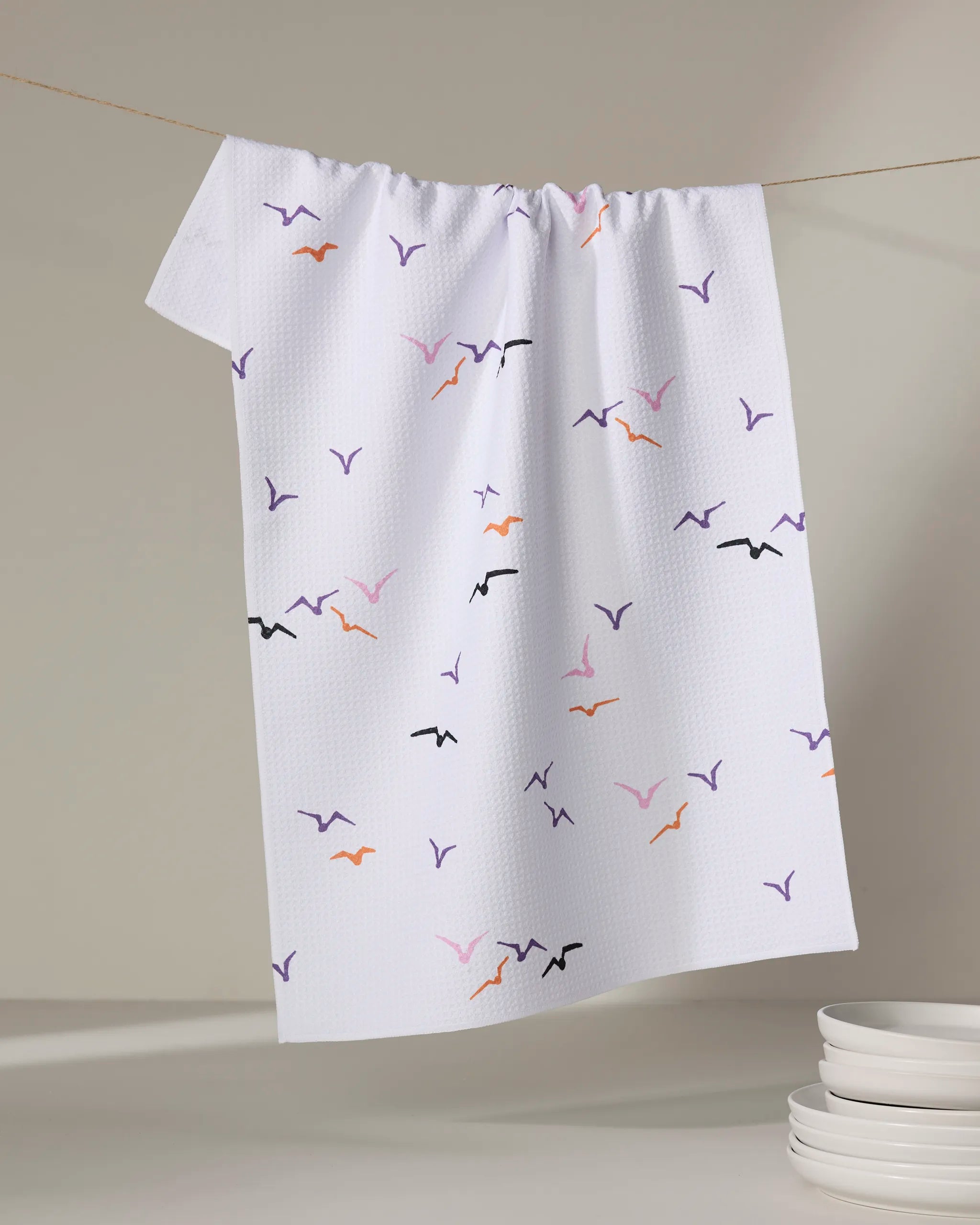 The "Scattered Seagulls" tea towel by Ania Zwara Art, featuring vibrant bird silhouettes, hangs on a line beside a stack of white dishes on a light surface.