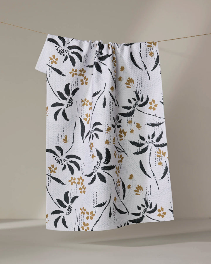 The Toboggan Avenue Scenic tea towel, featuring a quick-drying white fabric with a black and yellow floral pattern, hangs on a string against a neutral background.