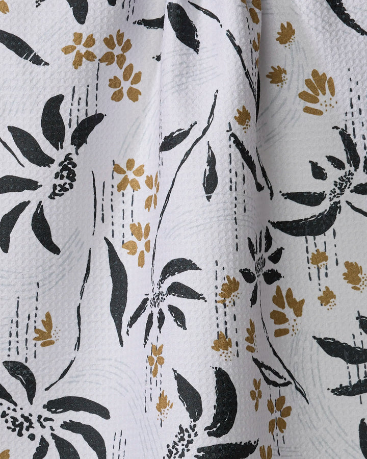 The Scenic tea towel by Toboggan Avenue features a textured white fabric with black floral shapes, mustard yellow flowers, fine dashed lines, and is made from quick-drying recycled materials.