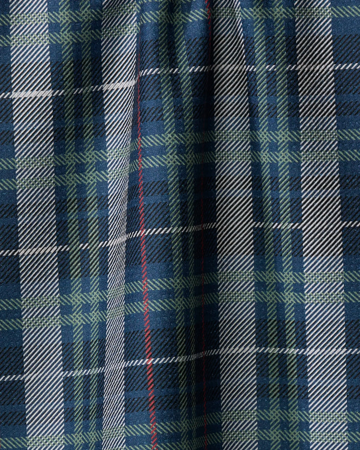 A close-up of Becki Owens' Scotty tea towel, featuring blue, green, black, and white tartan plaid with a thin red vertical stripe. The fabric is shown slightly pleated or draped.