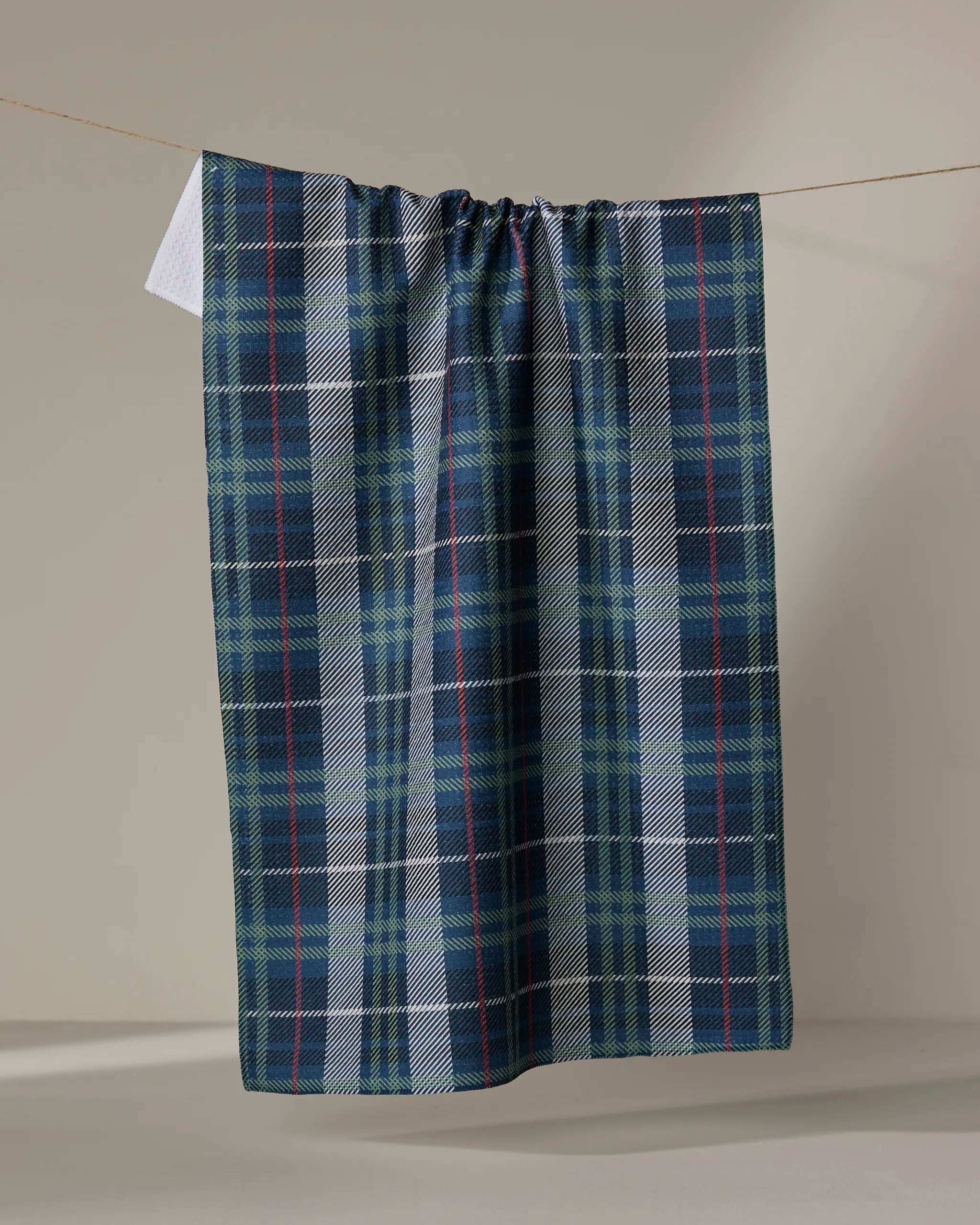 The Becki Owens Scotty kitchen towel, featuring blue, green, and white plaid, hangs on a clothesline against a plain beige background.