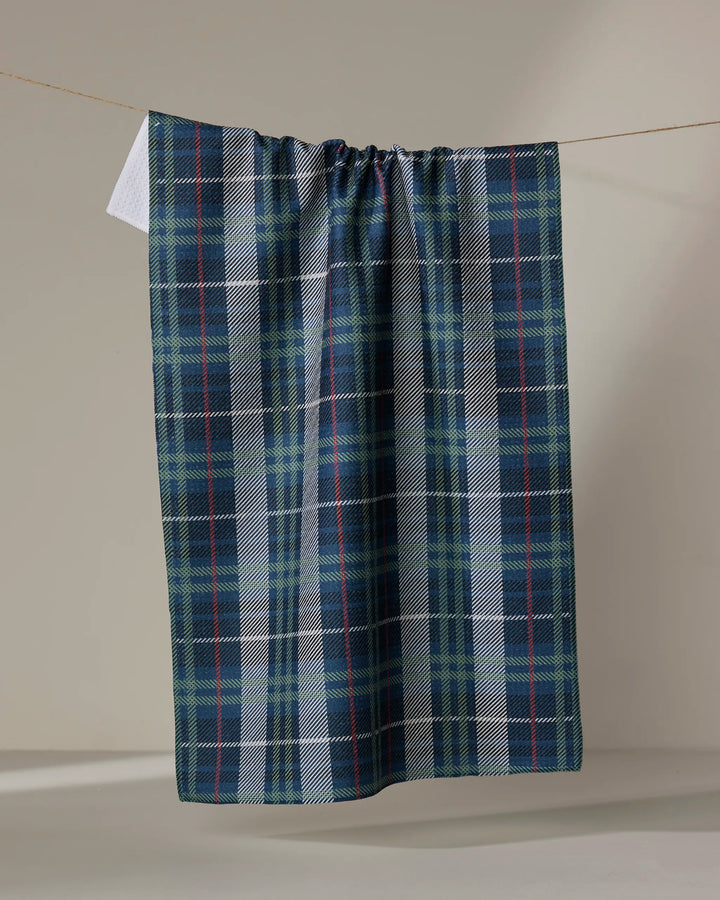 The Becki Owens Scotty kitchen towel, featuring blue, green, and white plaid, hangs on a clothesline against a plain beige background.