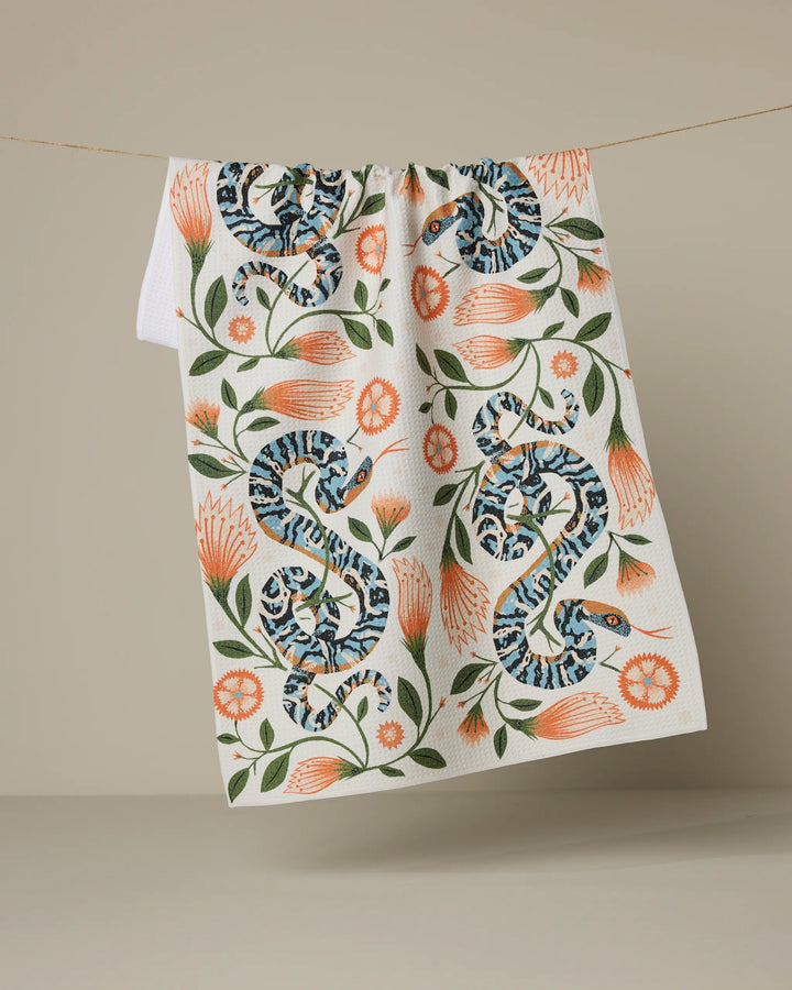 The Geometry "Scutes" tea towel features two blue patterned snakes and orange flowers with green leaves, printed on white fabric and shown hanging against a neutral beige background.