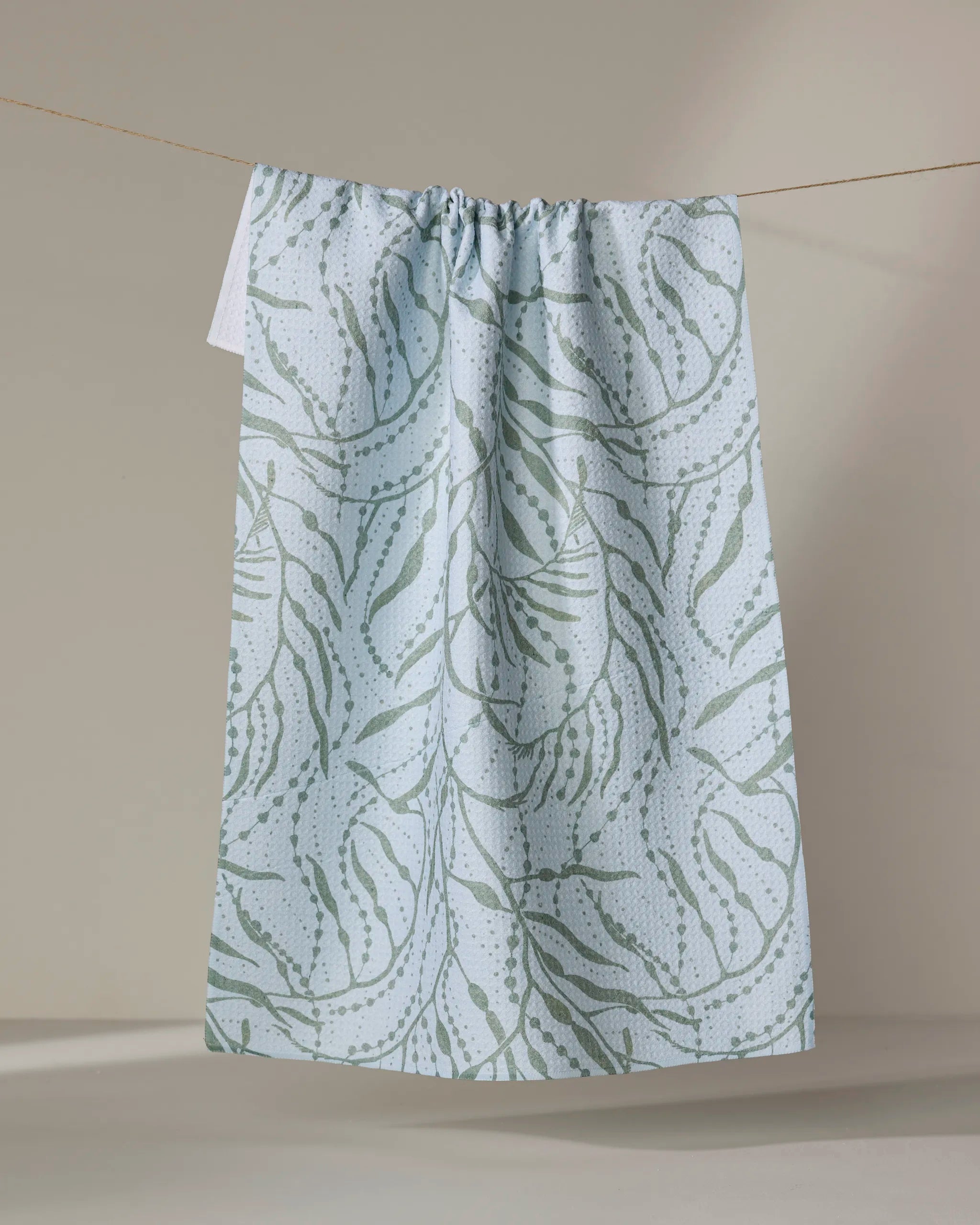Katie O'Shea's Sea Forest recycled kitchen towel, in pale blue with a subtle green botanical pattern, hangs on a thin clothesline against a neutral beige background.