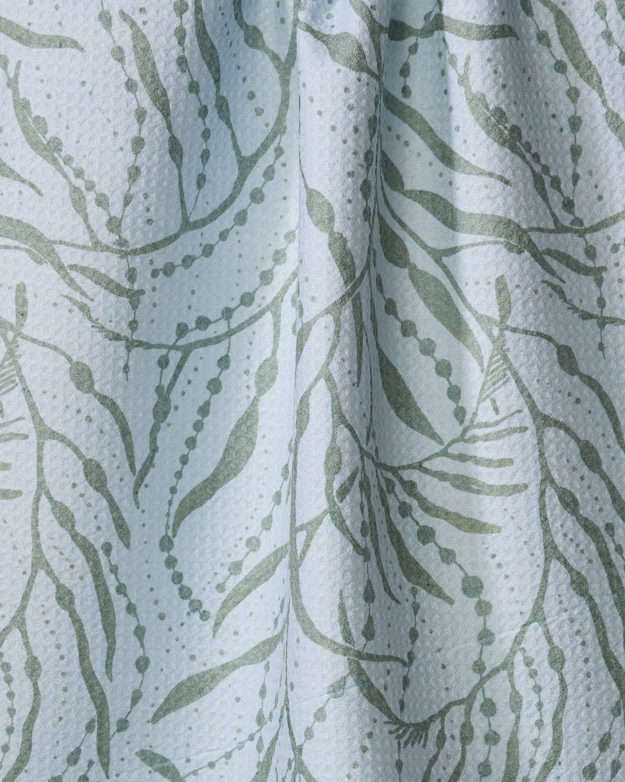 A close-up of Katie O’Shea’s Sea Forest shows textured white fabric with pale green leafy vines, small dots, and elongated leaf shapes. The slightly crumpled surface adds a charming, eco-friendly touch.