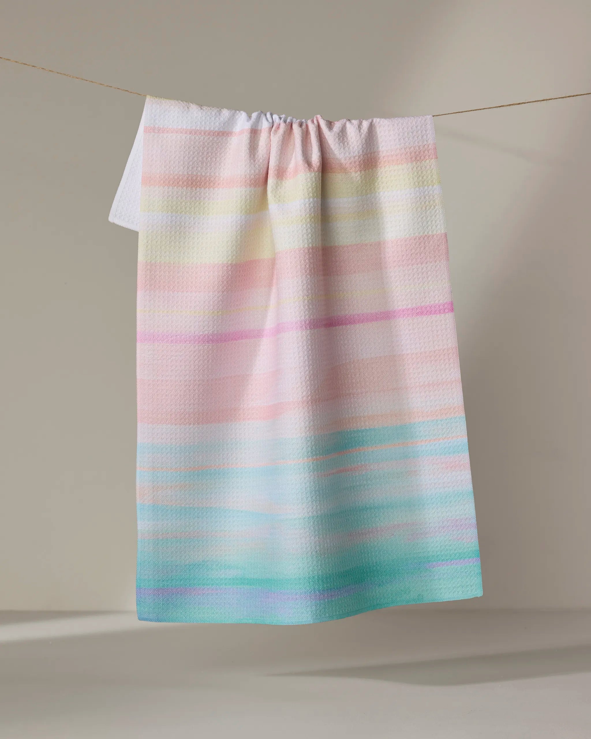 The Stefanie Bales Seafoam towel, featuring pastel horizontal stripes in pink, yellow, lavender, and aqua, hangs on a clothesline against a plain light background.