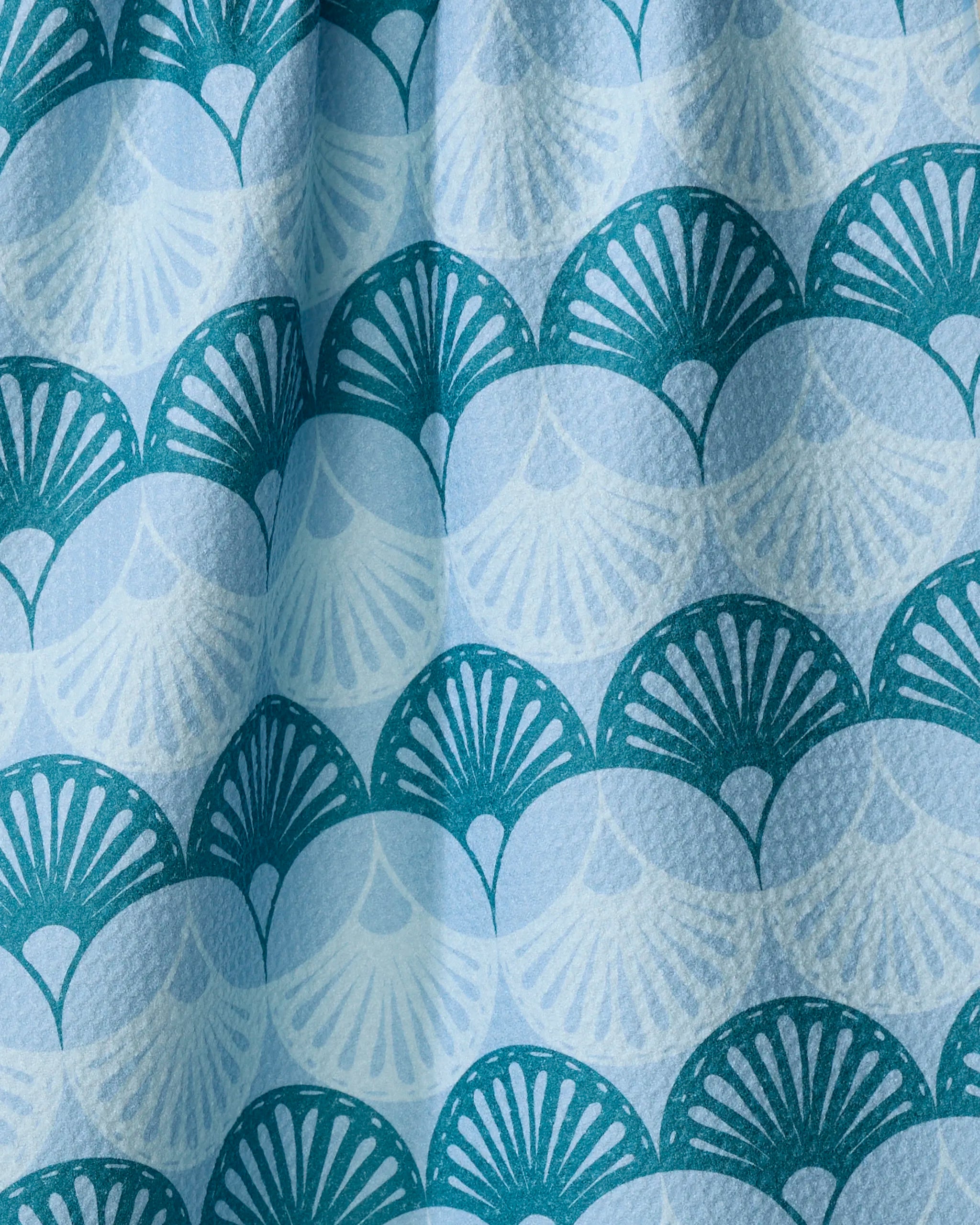 A close-up of the Marta Harvey "Seashells" eco-friendly kitchen towel, featuring a softly draped, textured fabric with a repeating blue and teal scallop pattern and fan-like designs.