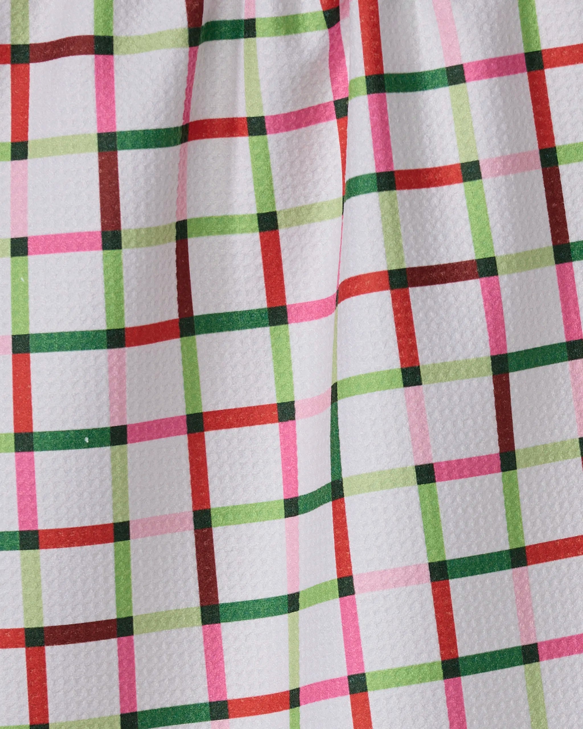A close-up of Geometry's Season of Joy recycled kitchen towel shows its textured white surface and colorful plaid pattern with green, red, and pink lines. The gathered fabric forms soft folds and shadows.