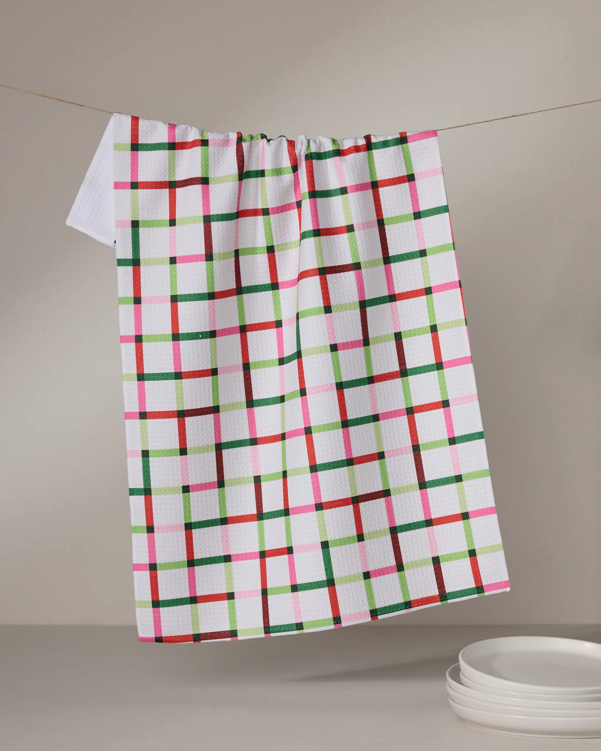 The Geometry Season of Joy tea towel, featuring a red, green, and pink checkered pattern, hangs above white plates and brings charm to any neutral kitchen with its quick-drying fabric.
