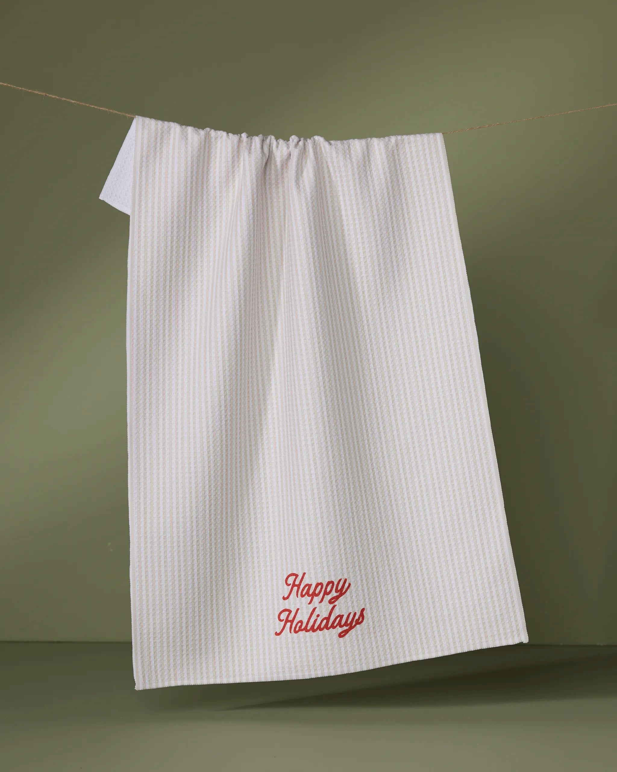 The Geometry Seasons Stripes tea towel, ultra-absorbent with subtle white stripes, hangs on a green backdrop. “Happy Holidays” is embroidered in red near the bottom of this festive kitchen essential.