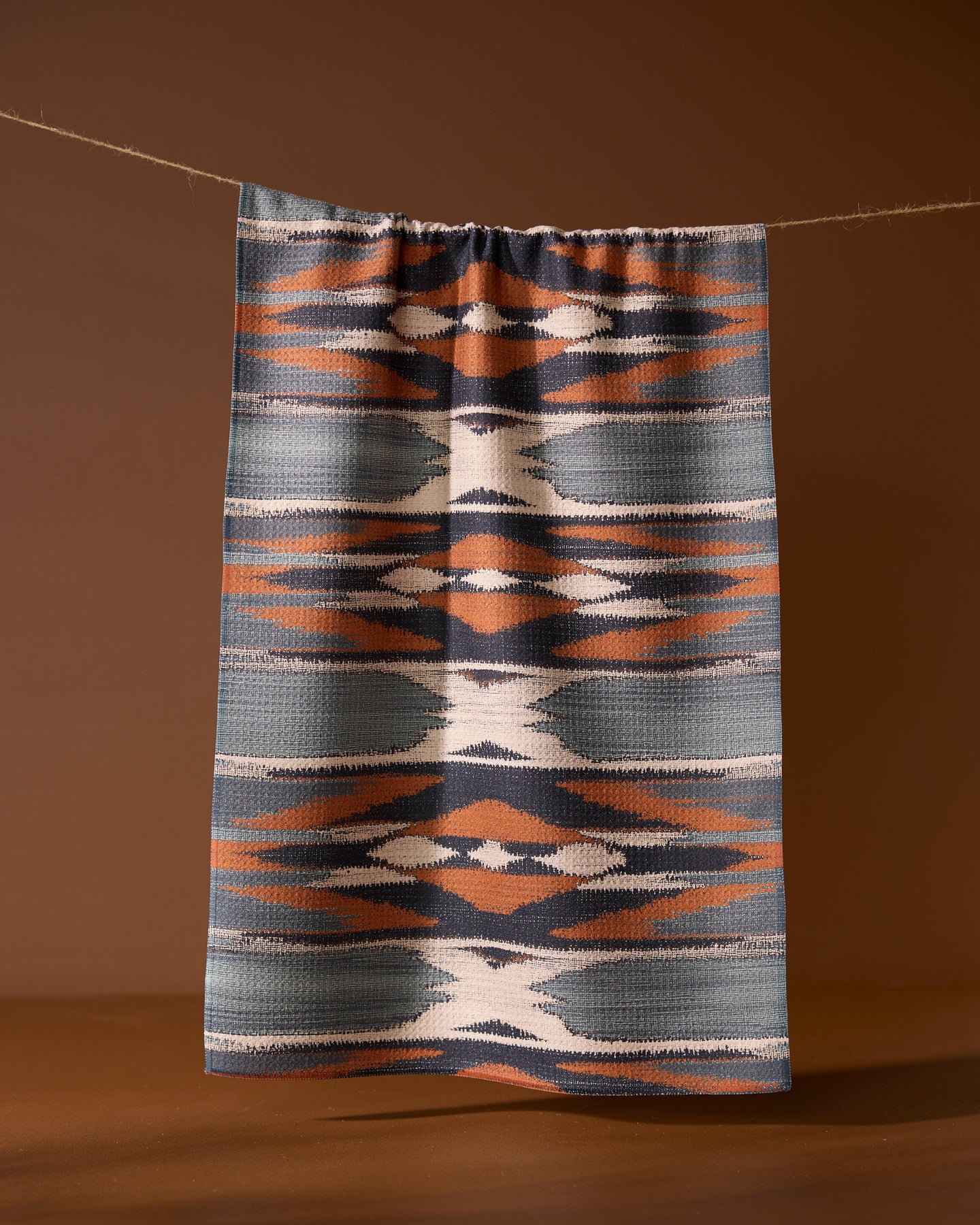 The Sedona kitchen towel by Geometry, featuring geometric blue, orange, white, and gray patterns, hangs from a string against a brown background.