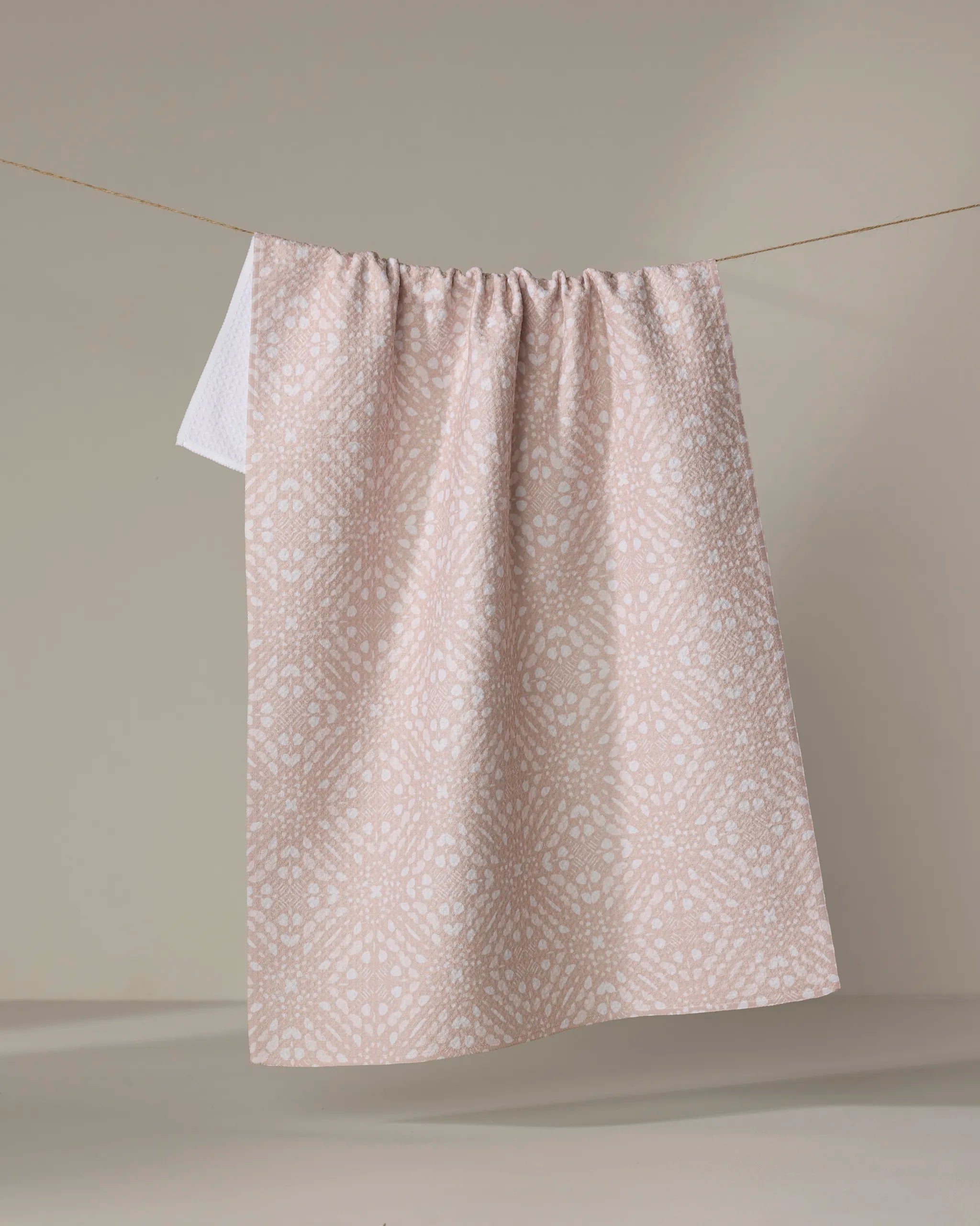 A Seeds and Nuts Neutral dish towel by Twig + Spark in light pink with a subtle white spotted pattern hangs neatly on a clothesline against a plain beige background.