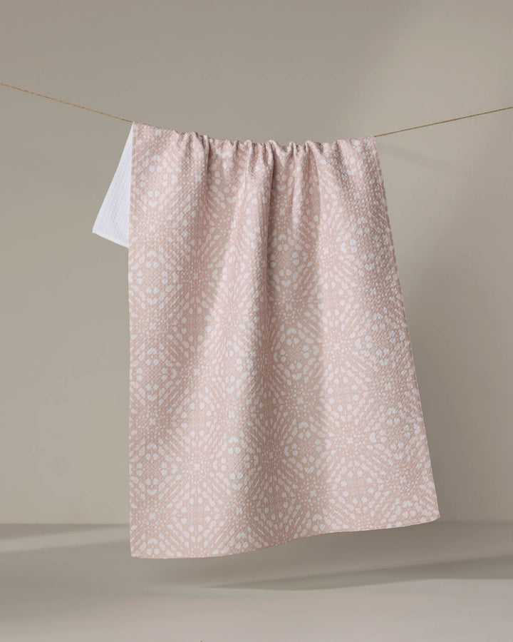 A Seeds and Nuts Neutral dish towel by Twig + Spark in light pink with a subtle white spotted pattern hangs neatly on a clothesline against a plain beige background.