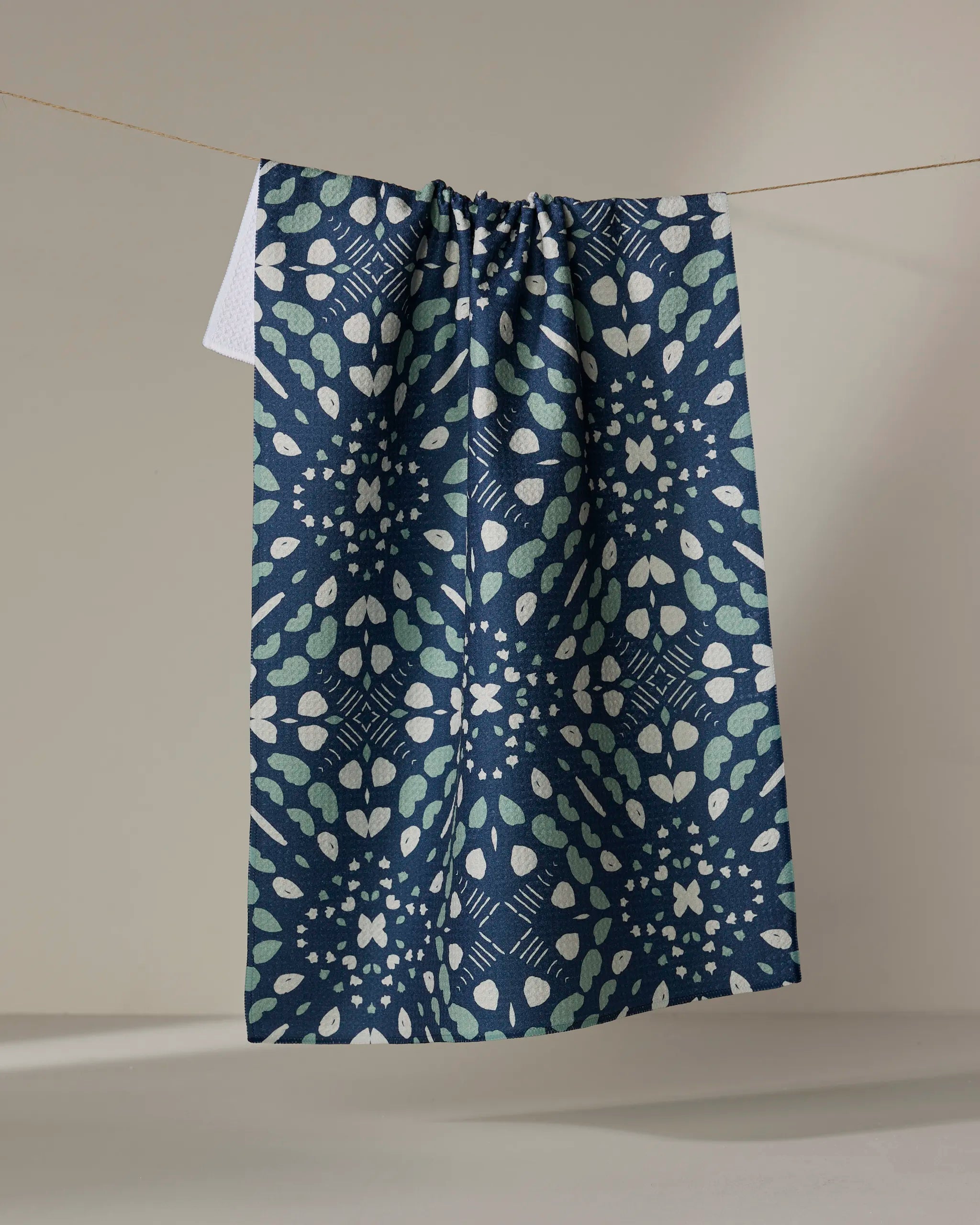 A Twig + Spark "Seeds and Nuts" blue dish towel with a white and green geometric pattern hangs on a clothesline against a light background.