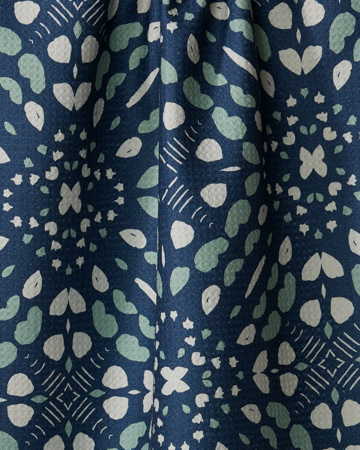 The Twig + Spark "Seeds and Nuts" kitchen towel features a dark blue eco-friendly fabric with a kaleidoscopic pattern of light green and white hearts, dots, and stars. This absorbent dish towel adds stylish charm and functionality to your kitchen.