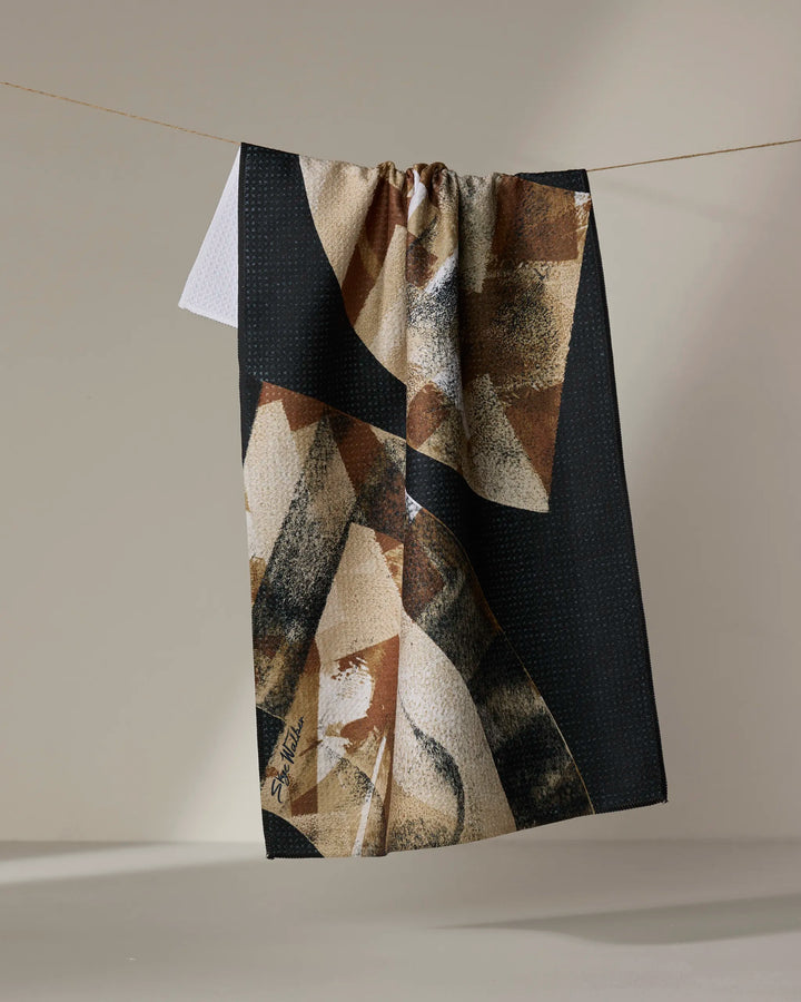 The Skye Walker Sepia Shift tea towel, featuring a geometric pattern in black, beige, and brown and made from recycled materials, hangs on a clothesline with part of a smaller white towel peeking out behind it.