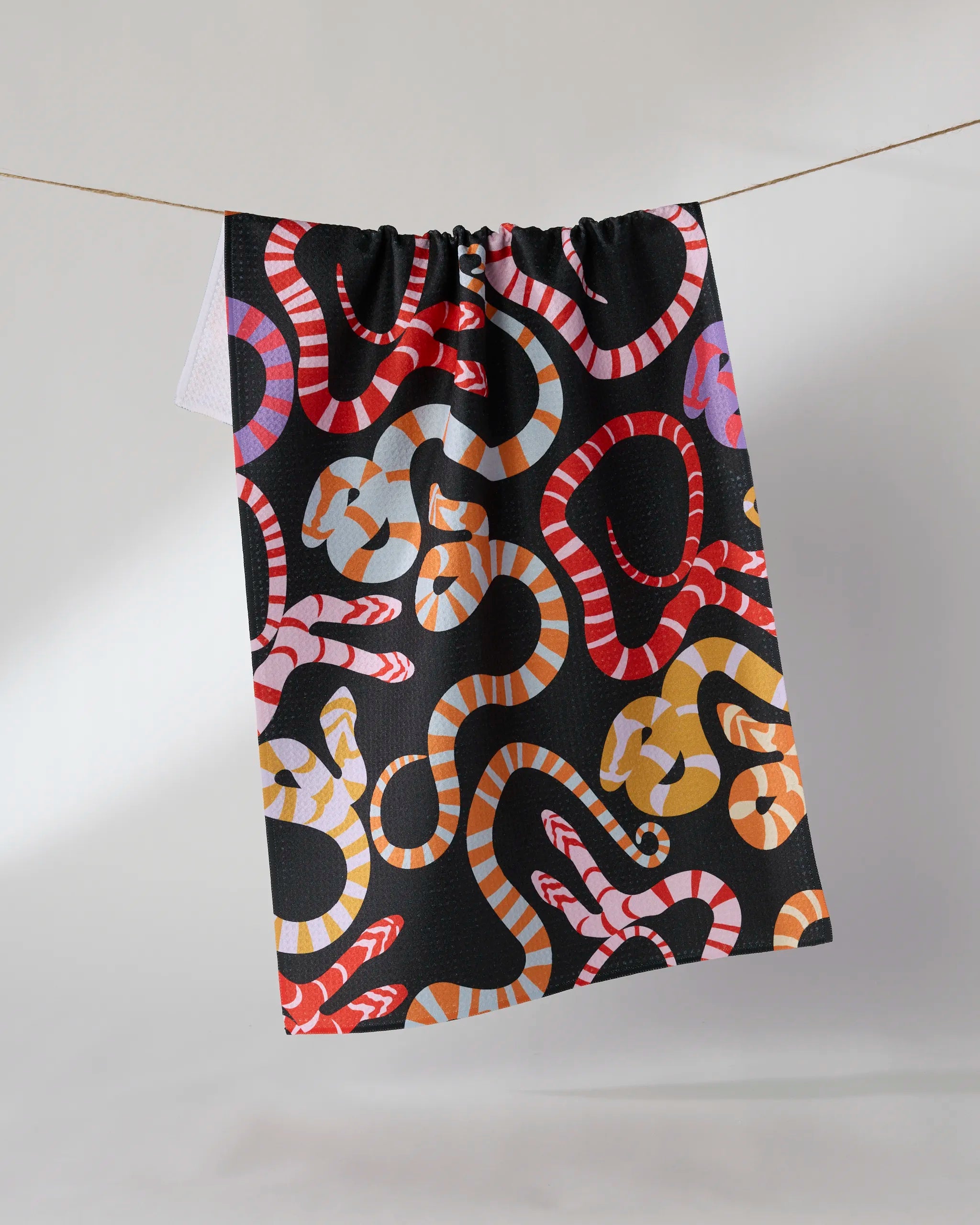 The Geometry Serpentine Riot towel, featuring a colorful red, orange, yellow, and white snake pattern on black, hangs on a line against a neutral background.