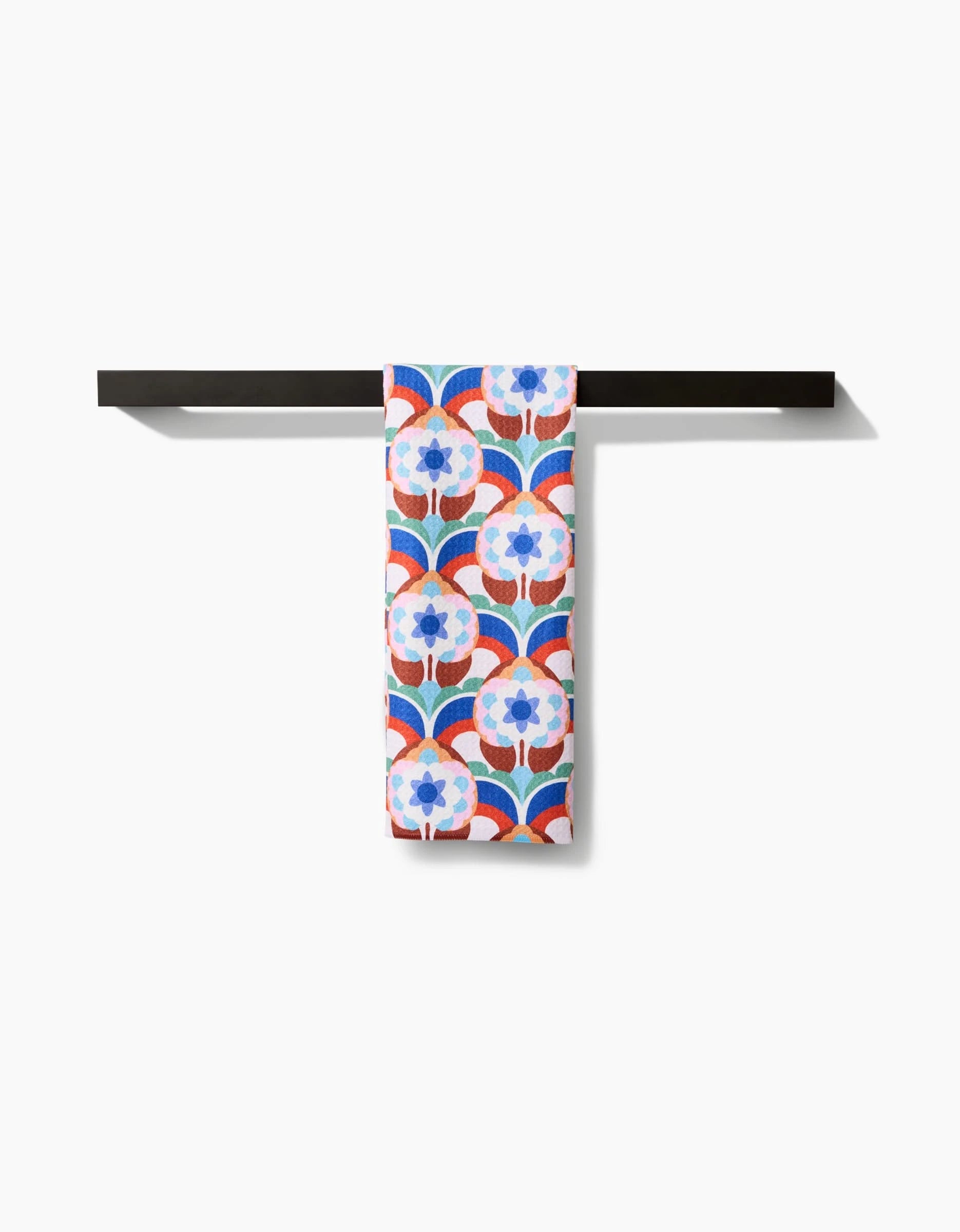 The Claire Iglesias Seventies Retro Flowers towel, ultra-absorbent with a blue, red, and white retro floral design, hangs neatly folded on a simple black towel bar against a white background.