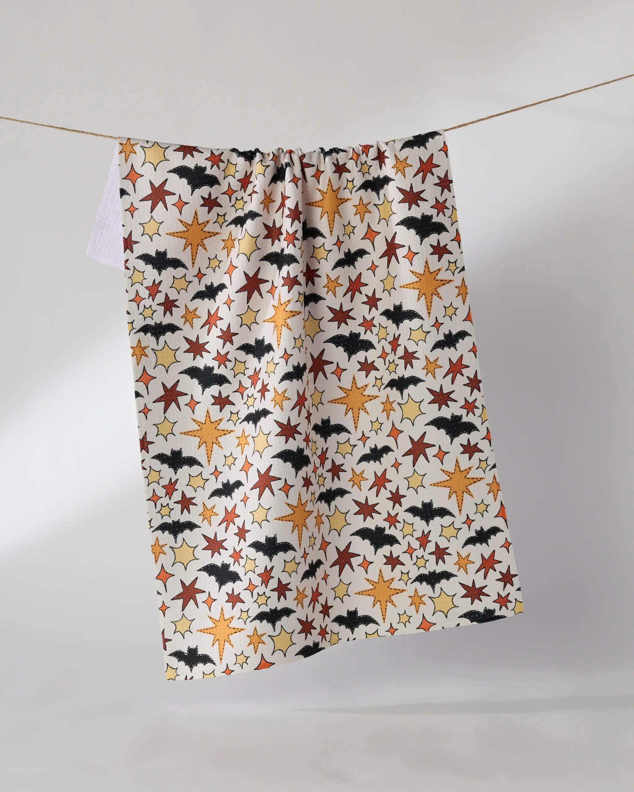 The Geometry Sew Batty tea towel, made from recycled materials, displays orange, red, and yellow stars with black bats and hangs on a clothesline against a plain white background.
