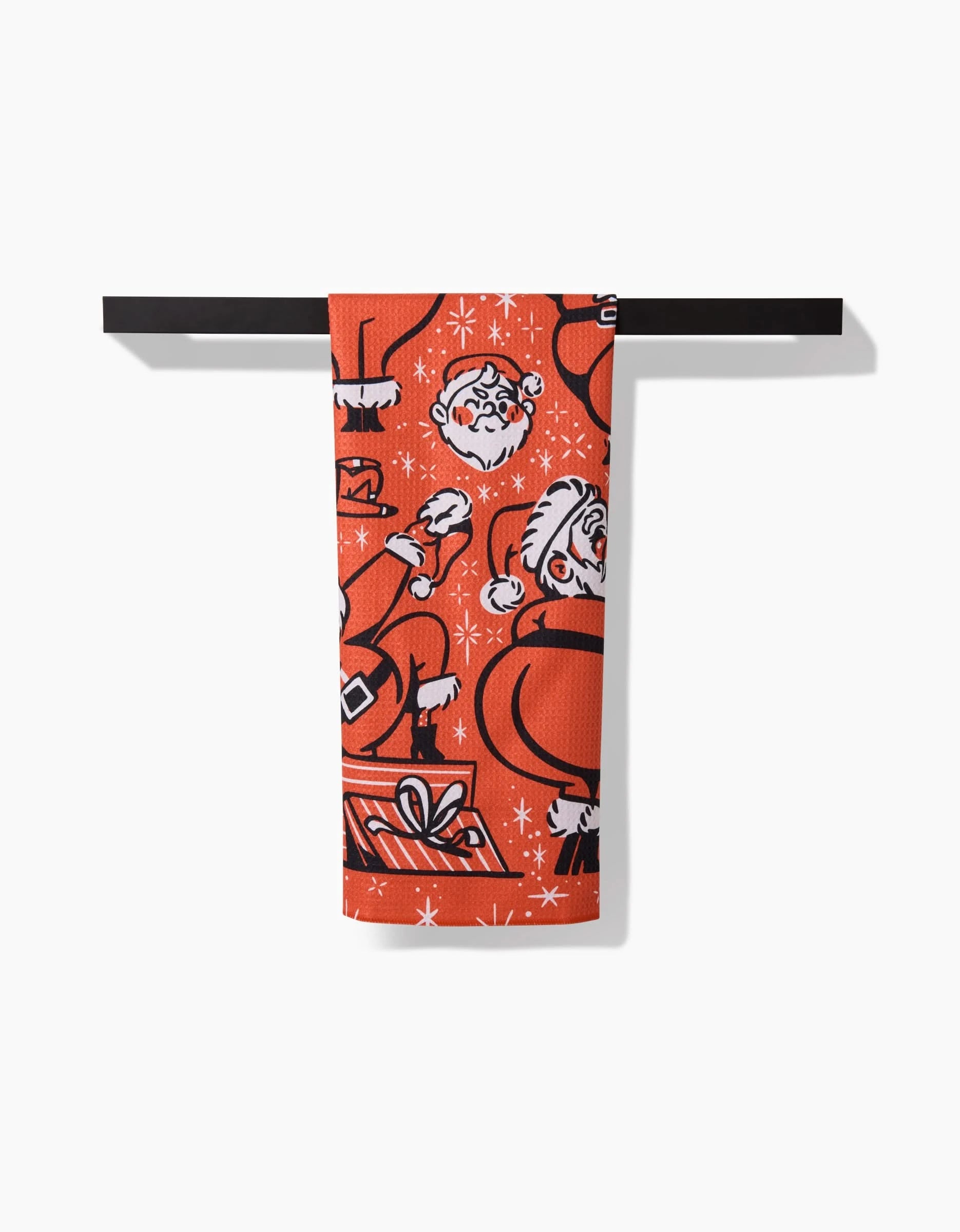 The Sexy Santa kitchen towel by Carly Berry is made from recycled materials, with red fabric and white and black Santa, gifts, candy canes, and snowflakes designs. It hangs on a black rod against a white background.