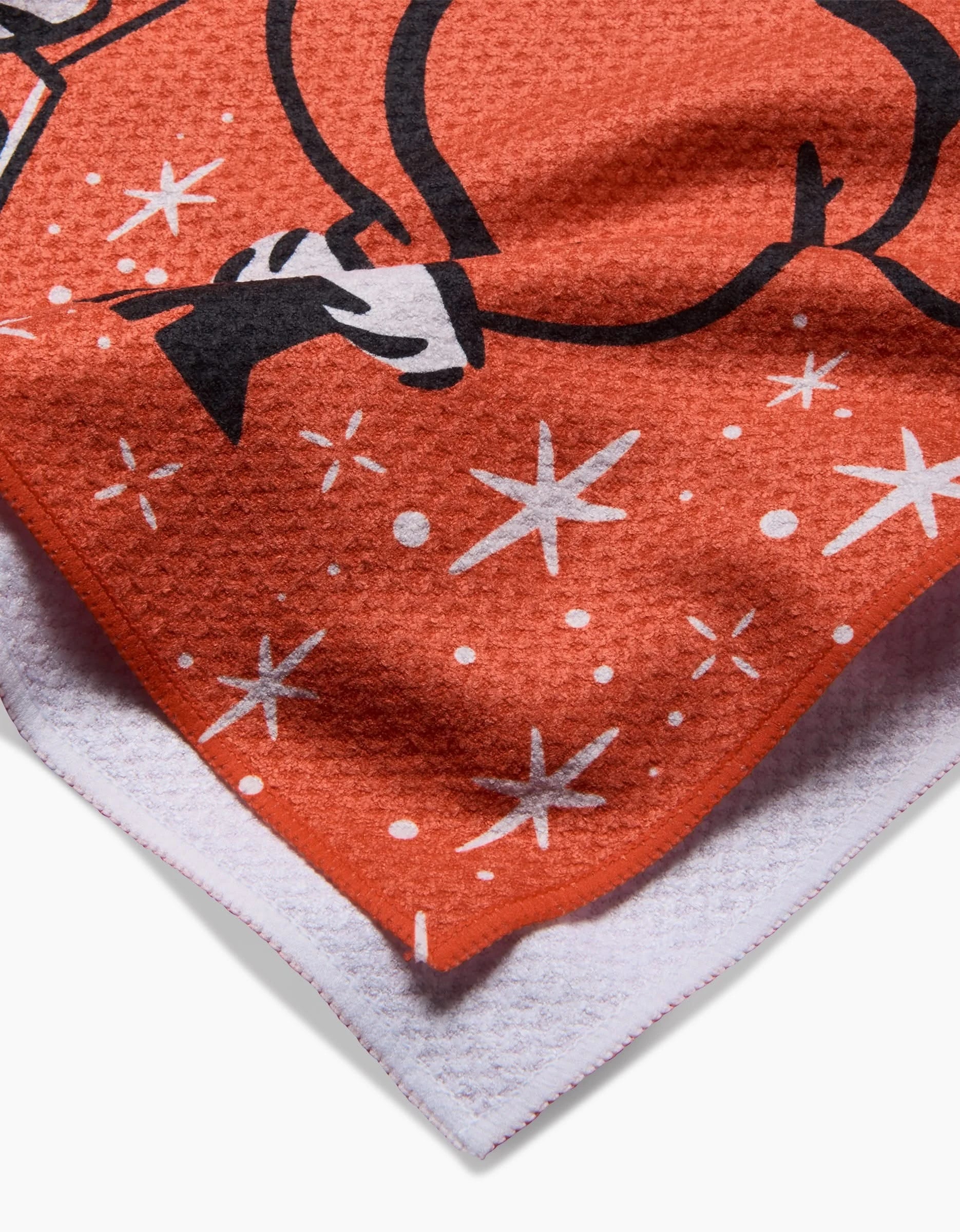 A close-up of Carly Berry’s Sexy Santa kitchen towel, featuring white stars, a black and white cartoon design, textured orange fabric, and white edges—crafted from recycled materials.