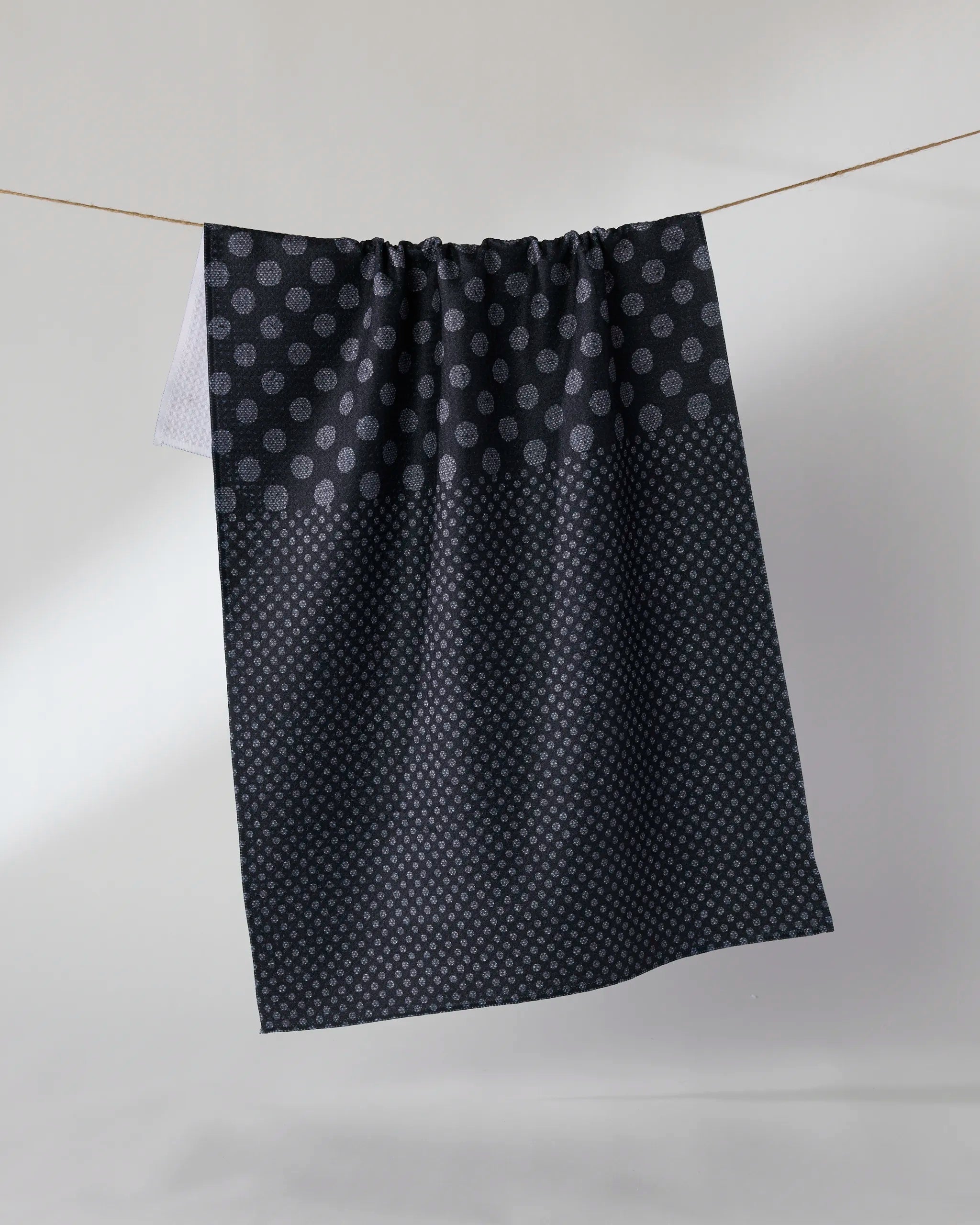 The Geometry Shadow Dot is a dark ultra-absorbent kitchen towel featuring small, light dots in two patterns, shown hanging from a string against a plain light background.