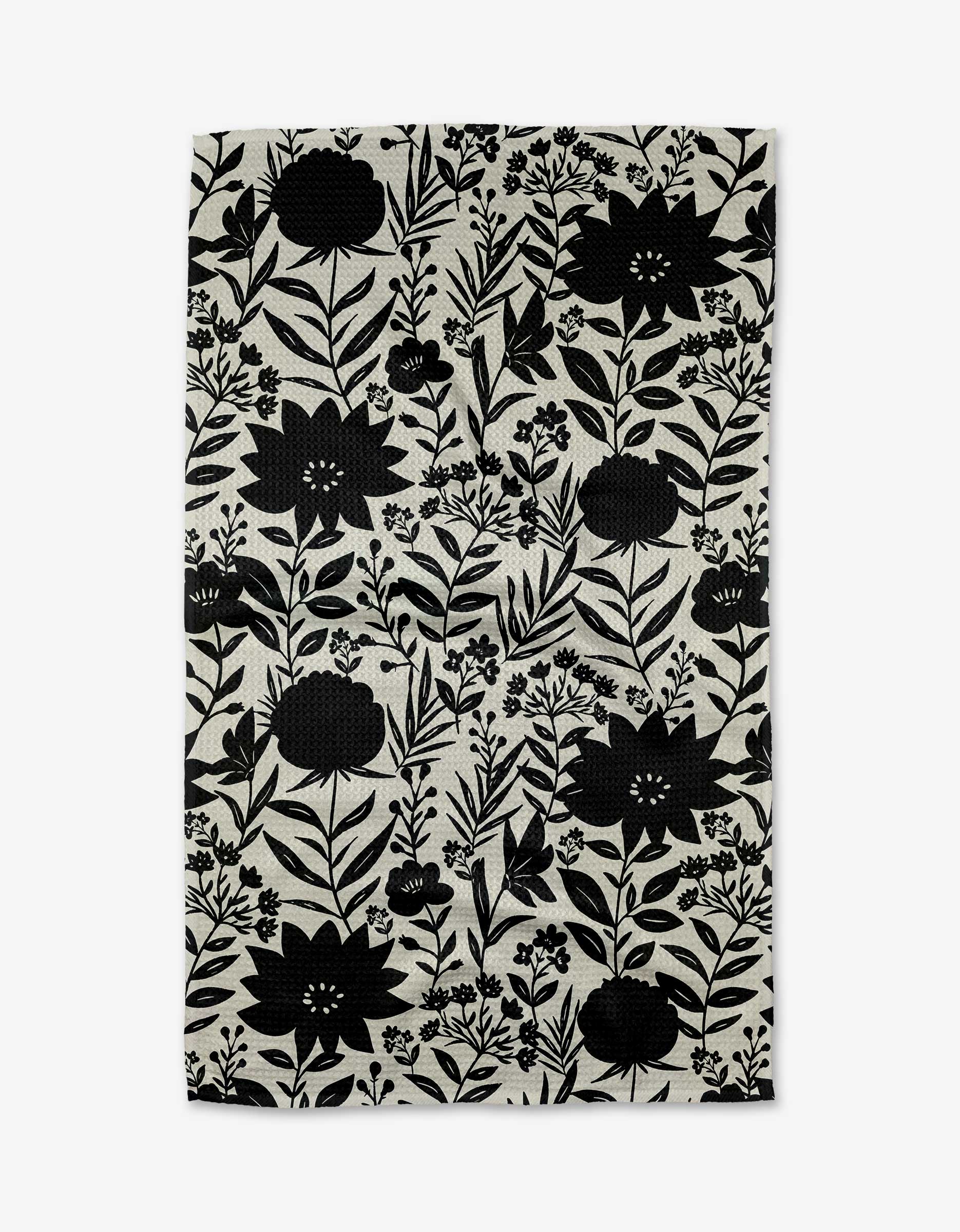 The Shadow Flower kitchen towel by Danelys Sidron features a rectangular, eco-friendly cloth adorned with a dense black floral and leafy pattern on a light beige background.