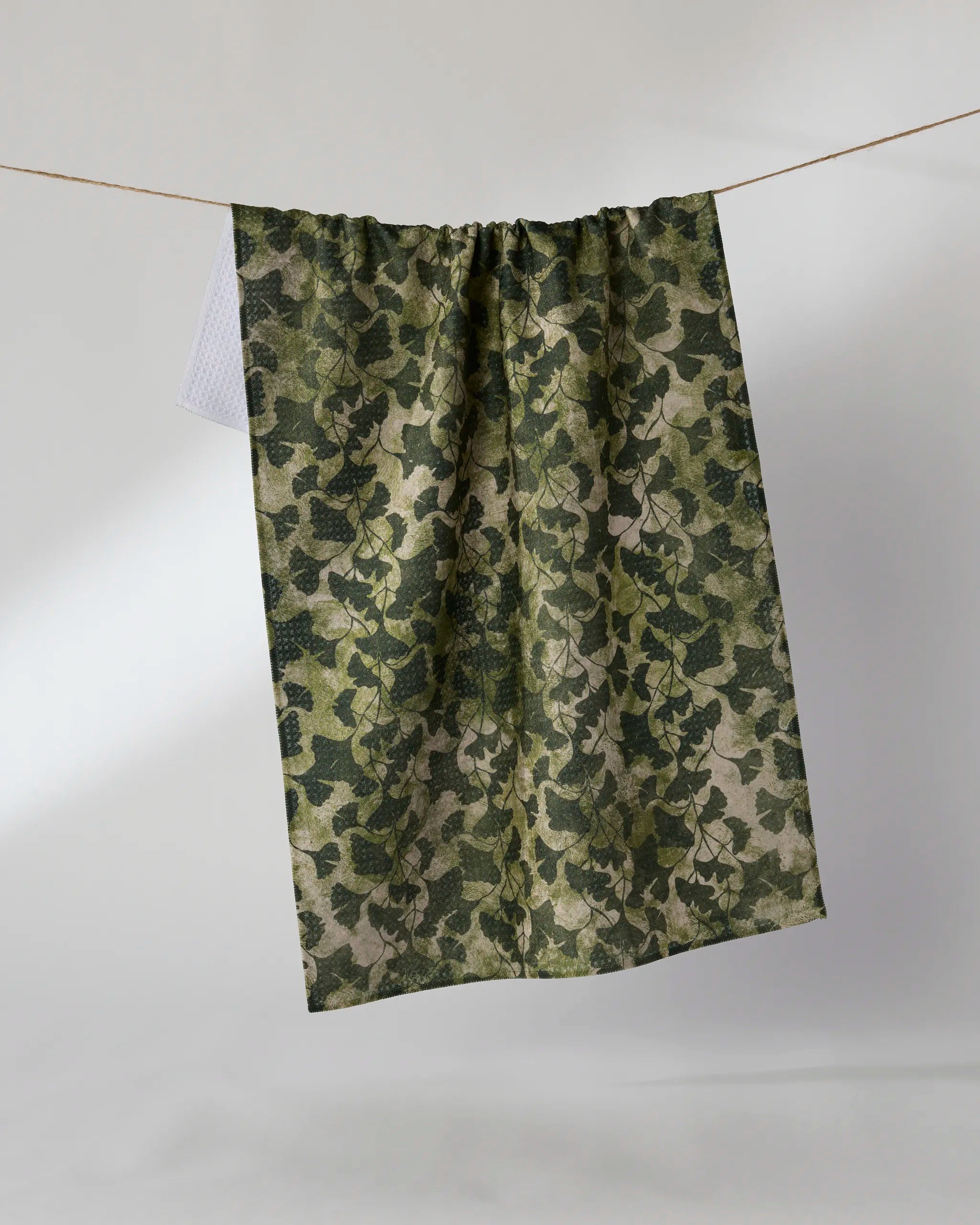 The Geometry Shadow Grove tea towel, featuring a green and beige ivy leaf pattern and made from recycled materials, hangs on a clothesline against a plain light background.