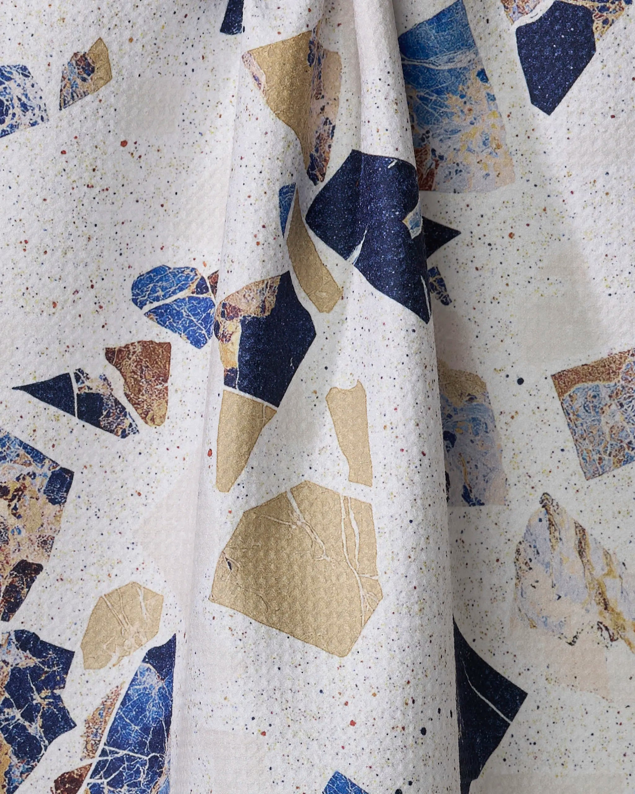 A close-up of Geometry’s Shale tea towel shows a terrazzo-inspired pattern with irregular blue, gold, and beige shapes on an off-white background with colorful speckles. The ultra-absorbent towel is slightly gathered in the center.