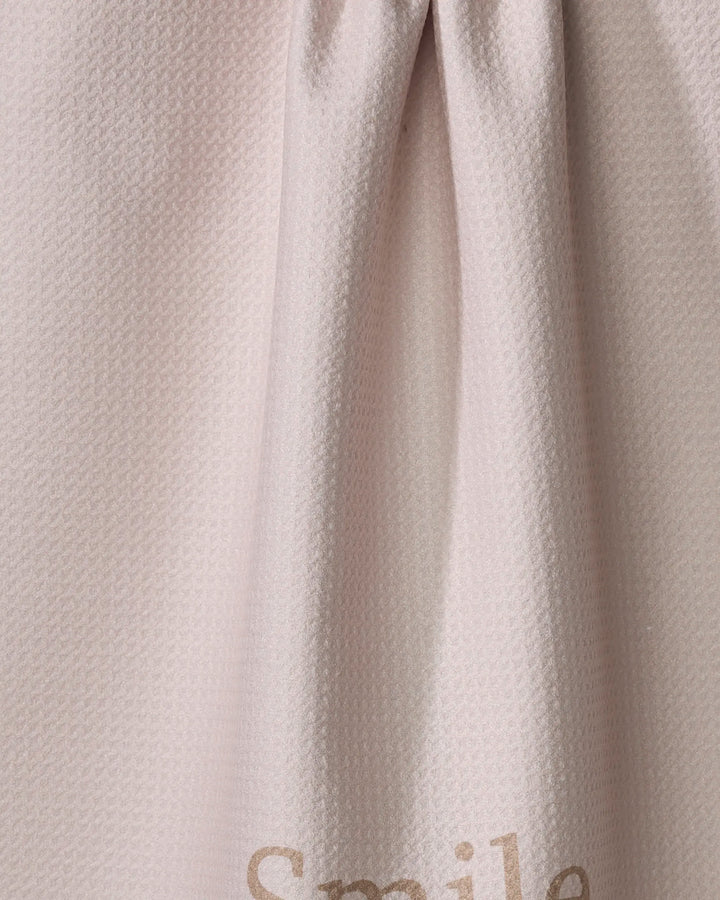 A close-up of Kelsi Fullmer's "Share A Smile" light beige kitchen tea towel shows subtle folds and the partial word “Smile” in light brown. Made from recycled materials, this towel is both stylish and sustainable.