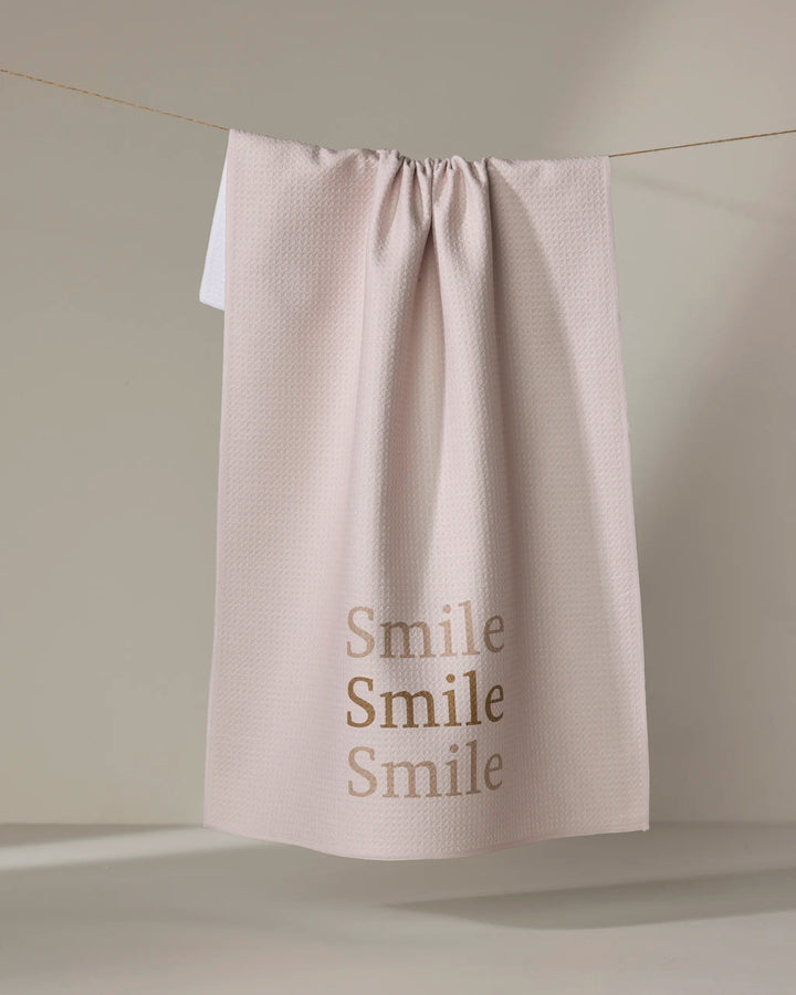 The Share A Smile kitchen tea towel by Kelsi Fullmer, in pale pink and made from recycled materials, hangs on a neutral background. “Smile” appears three times in gold lettering near the bottom for a cheerful touch.