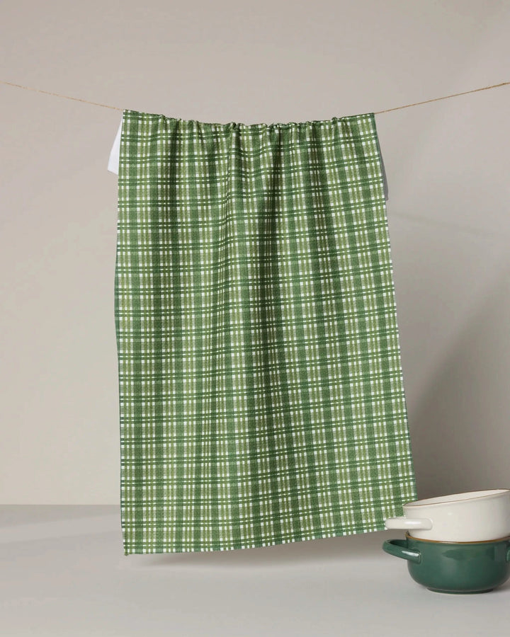 The Shay Plaid ultra-absorbent towel by Geometry, in green and white plaid, hangs on a string above a white table, with a matching green and white ceramic pot positioned below it on the right side.