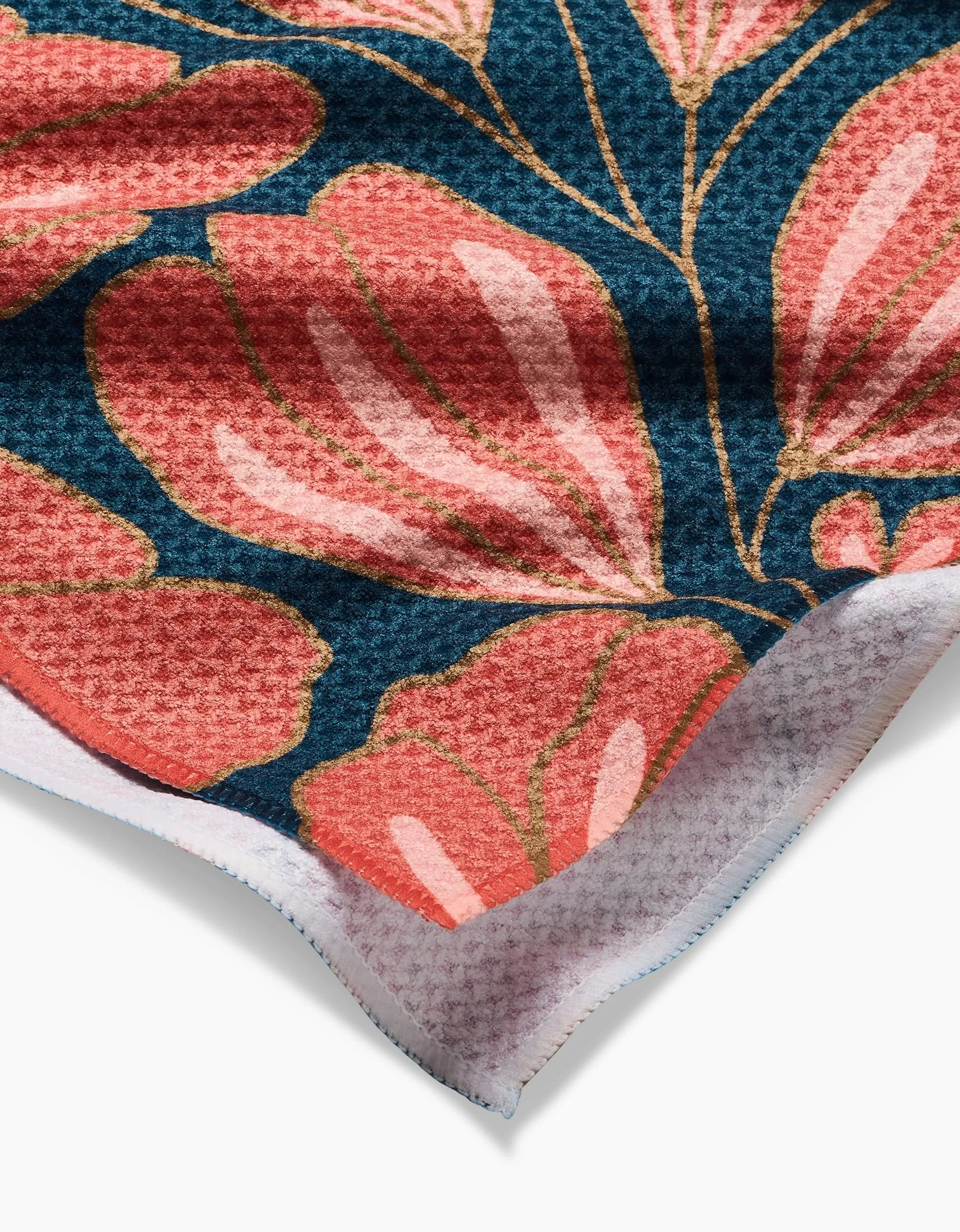 A close-up of the Sheila tea towel by Amy MacCready, made from recycled materials with a textured surface and bold floral design—large pink and red petals outlined in green on dark blue. A folded corner reveals the absorbent underside.
