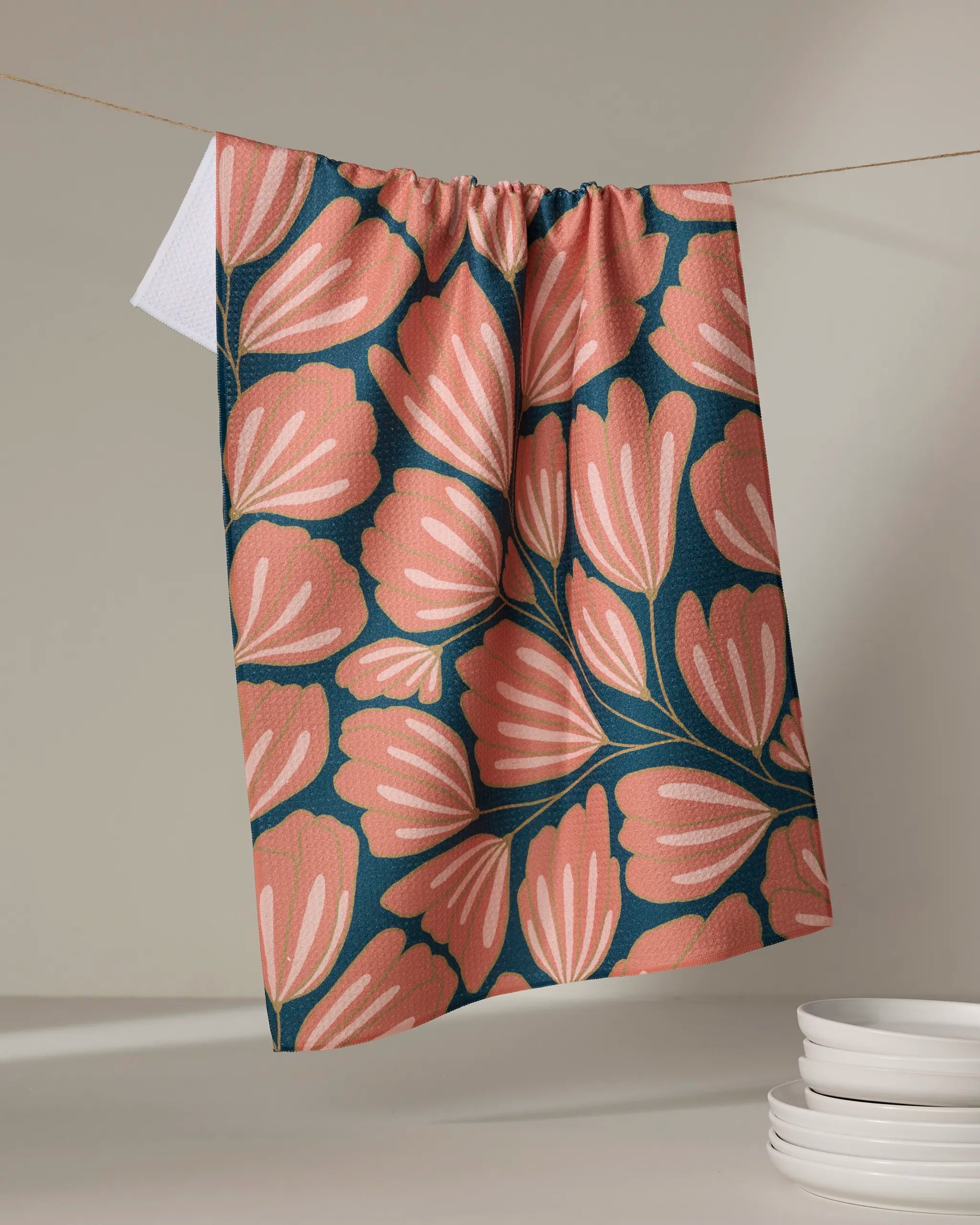The Sheila recycled kitchen towel by Amy MacCready, with bold pink and coral leaves on a dark teal background, hangs on a string above a stack of white plates.