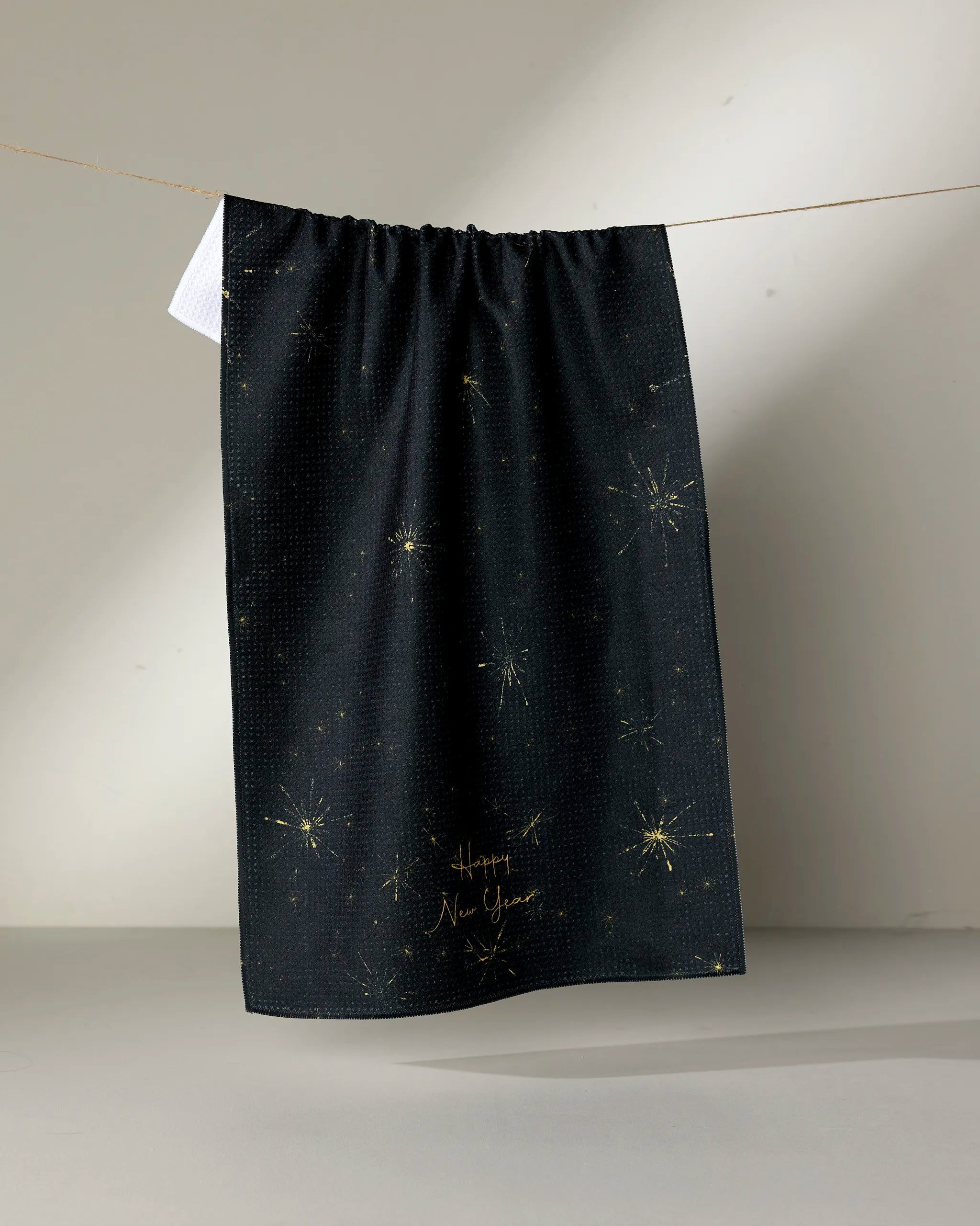 The Shimmer Eve kitchen towel by Geometry, made from recycled materials, displays gold starburst patterns and “Happy New Year” in gold on black fabric, hanging on a clothesline against a light, neutral background.